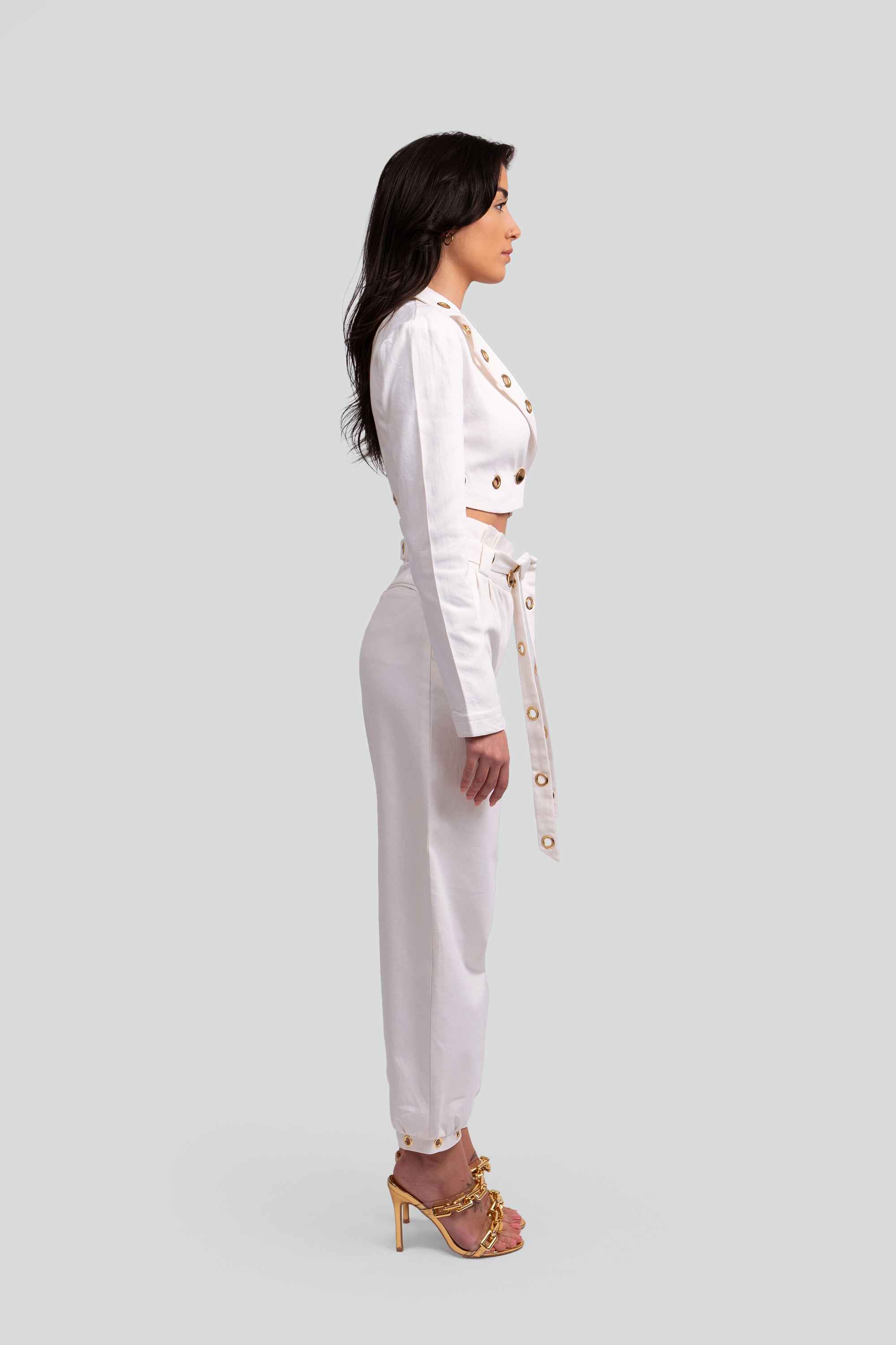 Side view of a woman wearing a white two piece cropped set with gold grommet details on a gray background