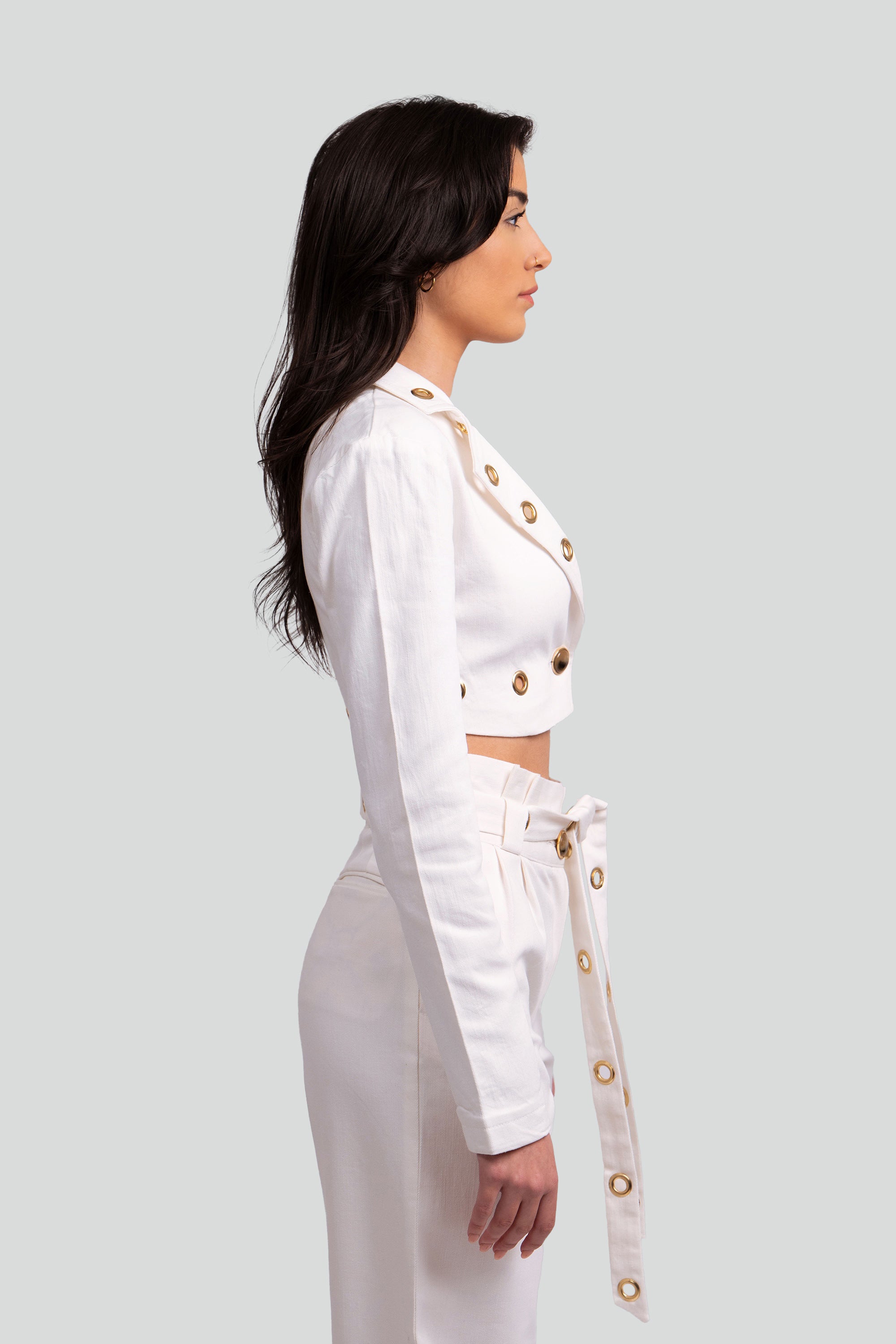 Side view of a woman wearing a white two piece cropped set with gold grommet details on a gray background