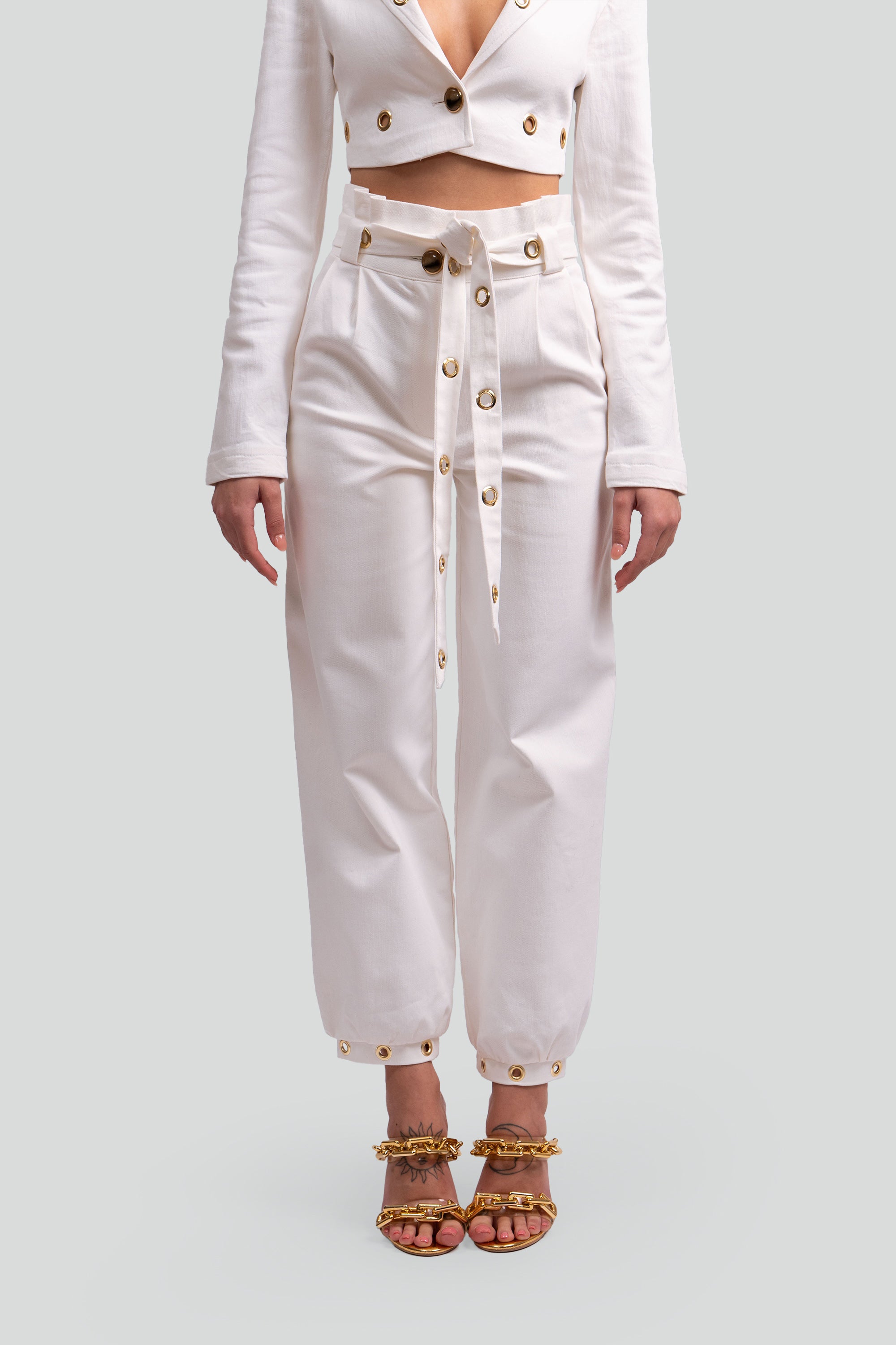 Close up view of a woman wearing a white two piece cropped set with gold grommet details on a gray background