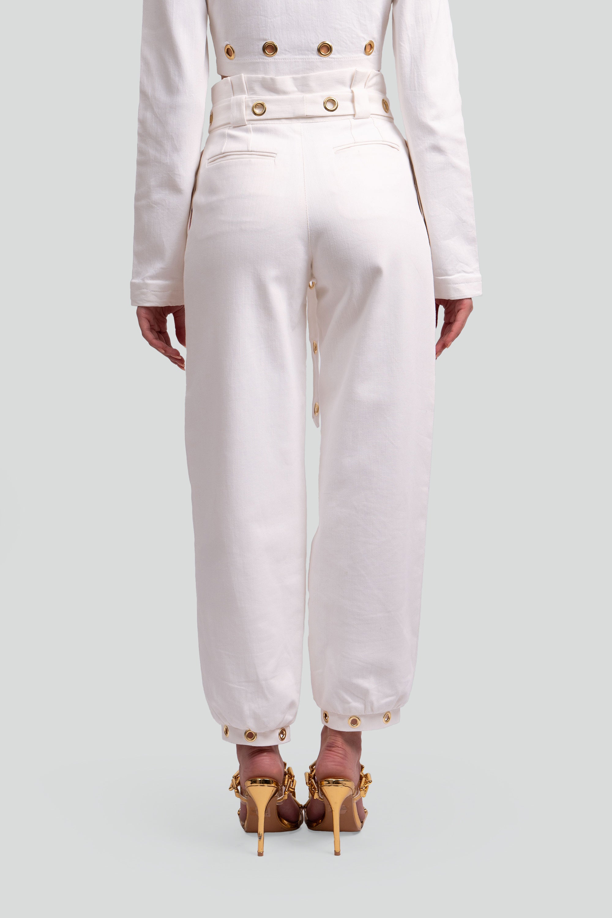 Back view of a woman wearing a white pant with gold grommet details on a gray background