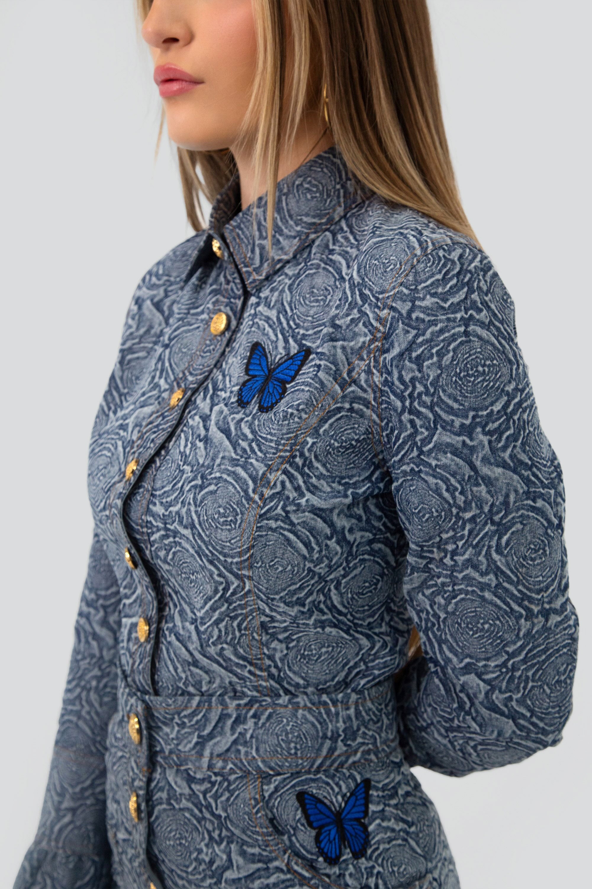 Woman wearing a blue patterned button up top and skirt set with butterfly embroidery on a plain background