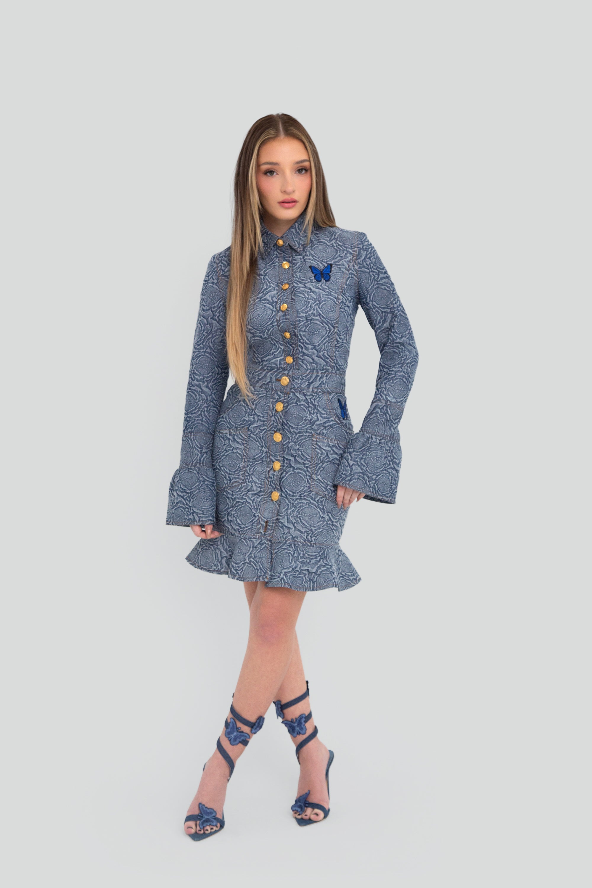 Woman wearing a blue patterned button up top and skirt set with flared sleeves on a plain background