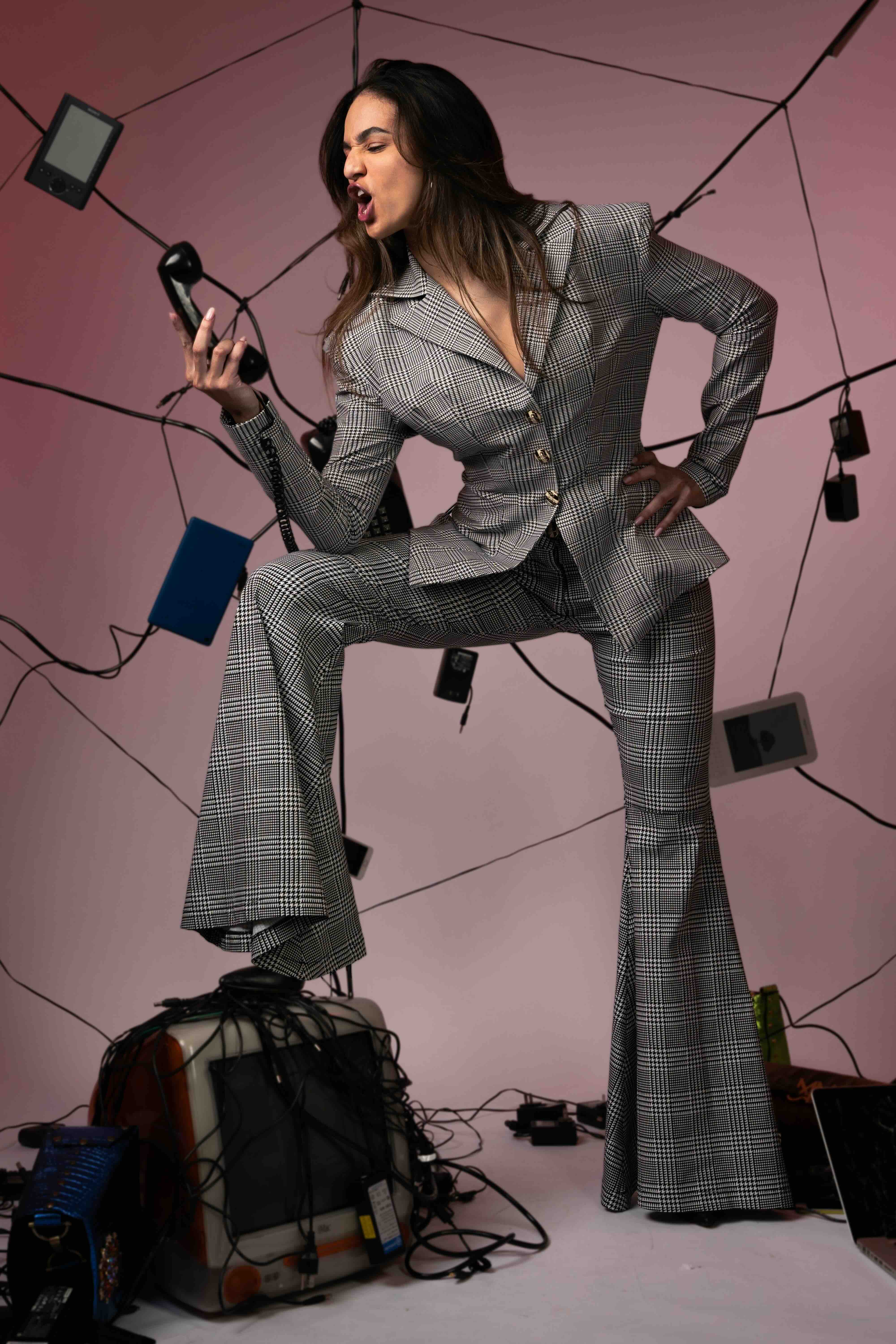 Woman in a grey plaid suit with one leg on a computer and phone in hand with electronic equipment around her against a pink background