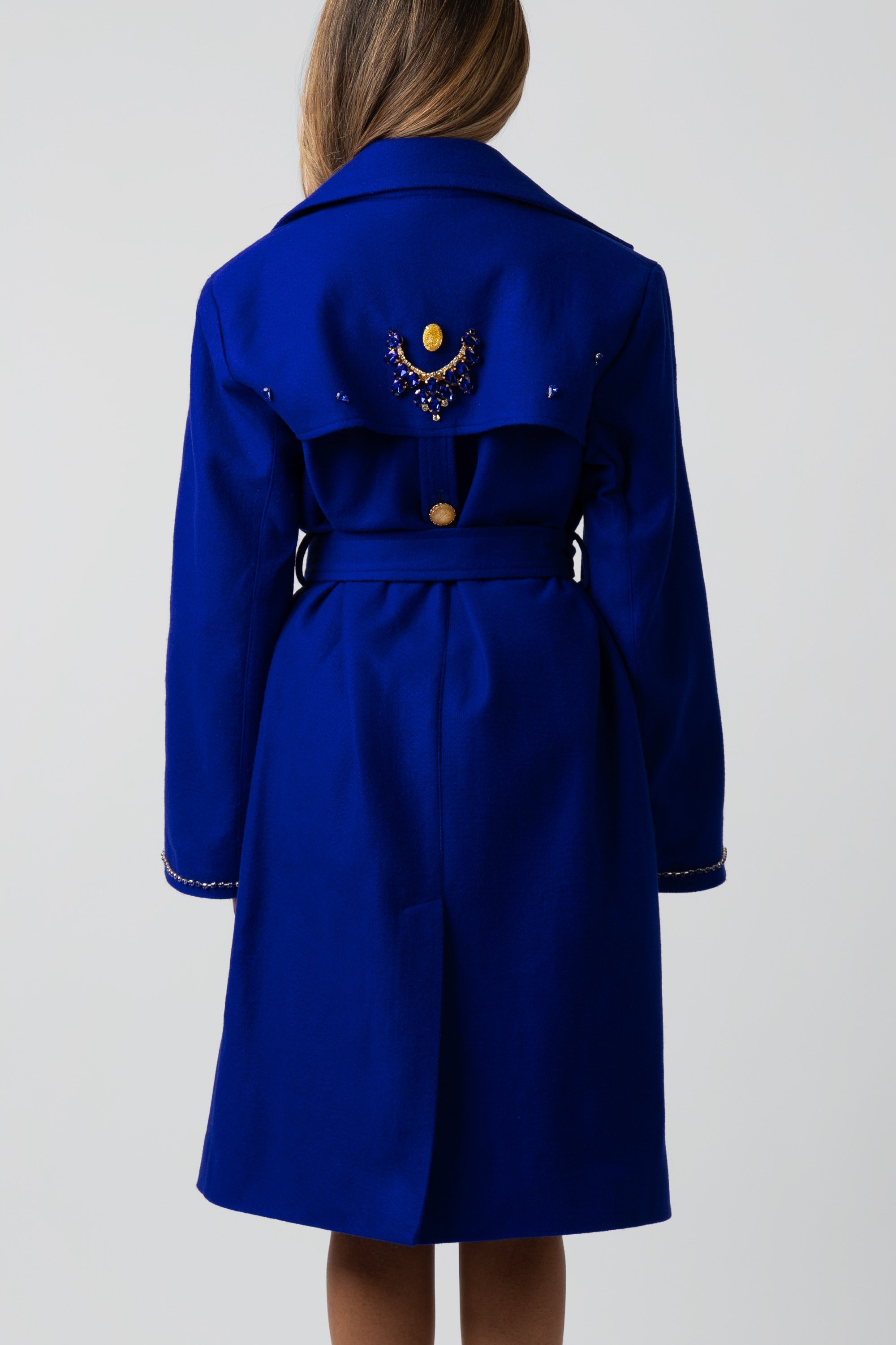 Back of a woman wearing a blue trench coat with gold detailing on a white background