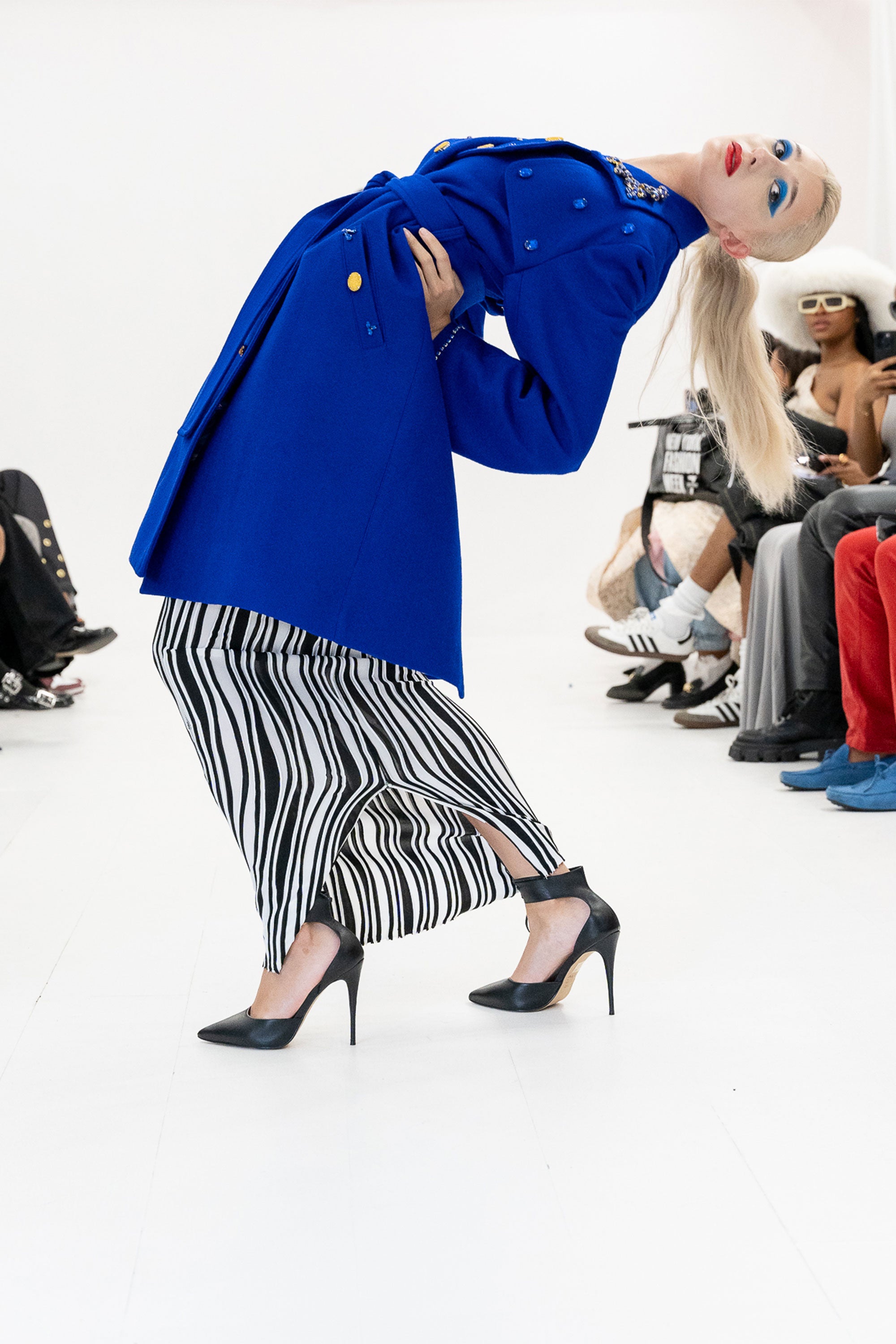 Model wearing a blue trench coat with gold detailing and black and white skirt posing against a white background and an audience