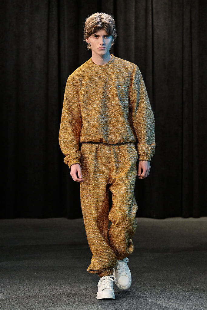 Model wearing a gold textured sweatsuit with white sneakers on a runway.