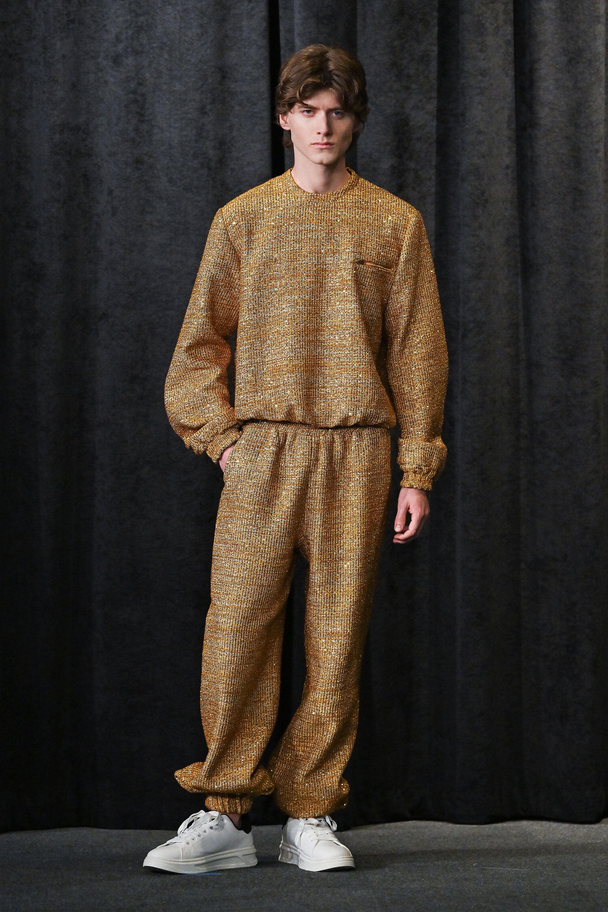 Person wearing a gold textured sweatsuit against a black curtain background