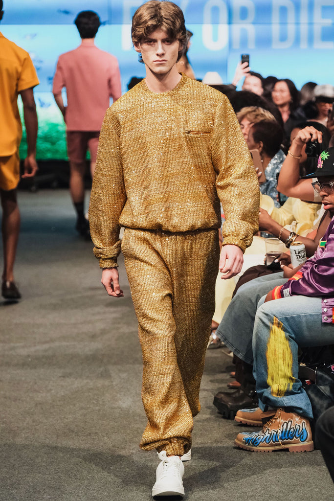 Model walking on a fashion runway wearing a gold textured sweatsuit.