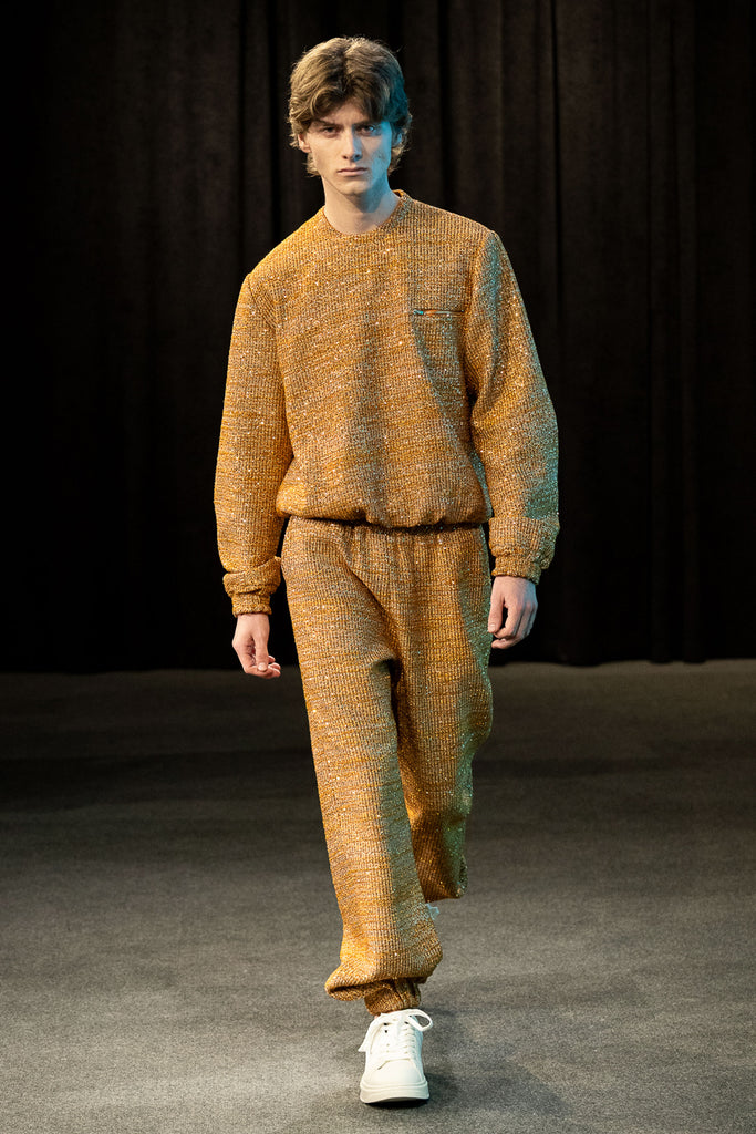 Model wearing a gold textured sweatsuit on a runway with a dark curtain background