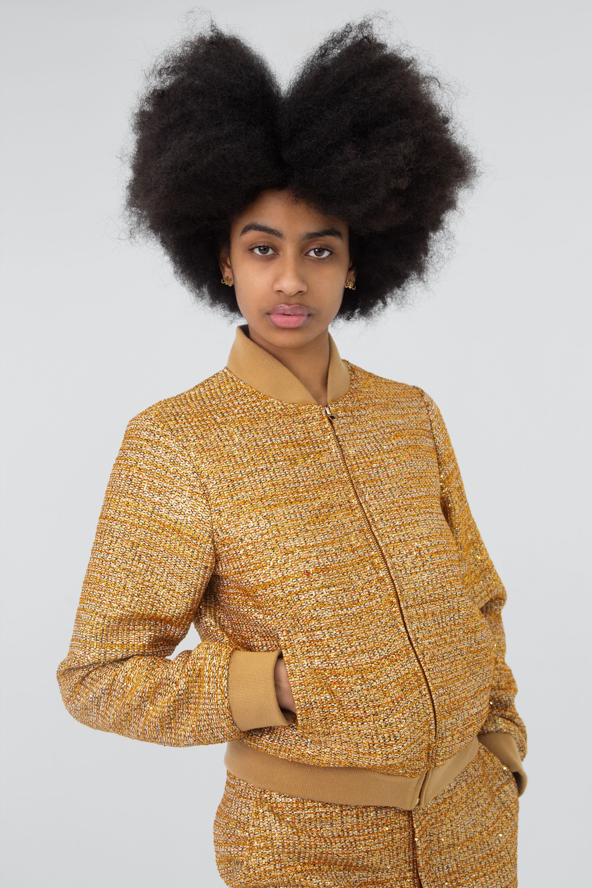 Close up of a woman wearing a gold textured tracksuit on a plain background