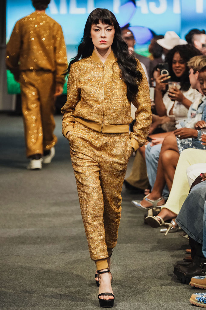 Model in a gold textured tracksuit walking on a runway with an audience in the background.
