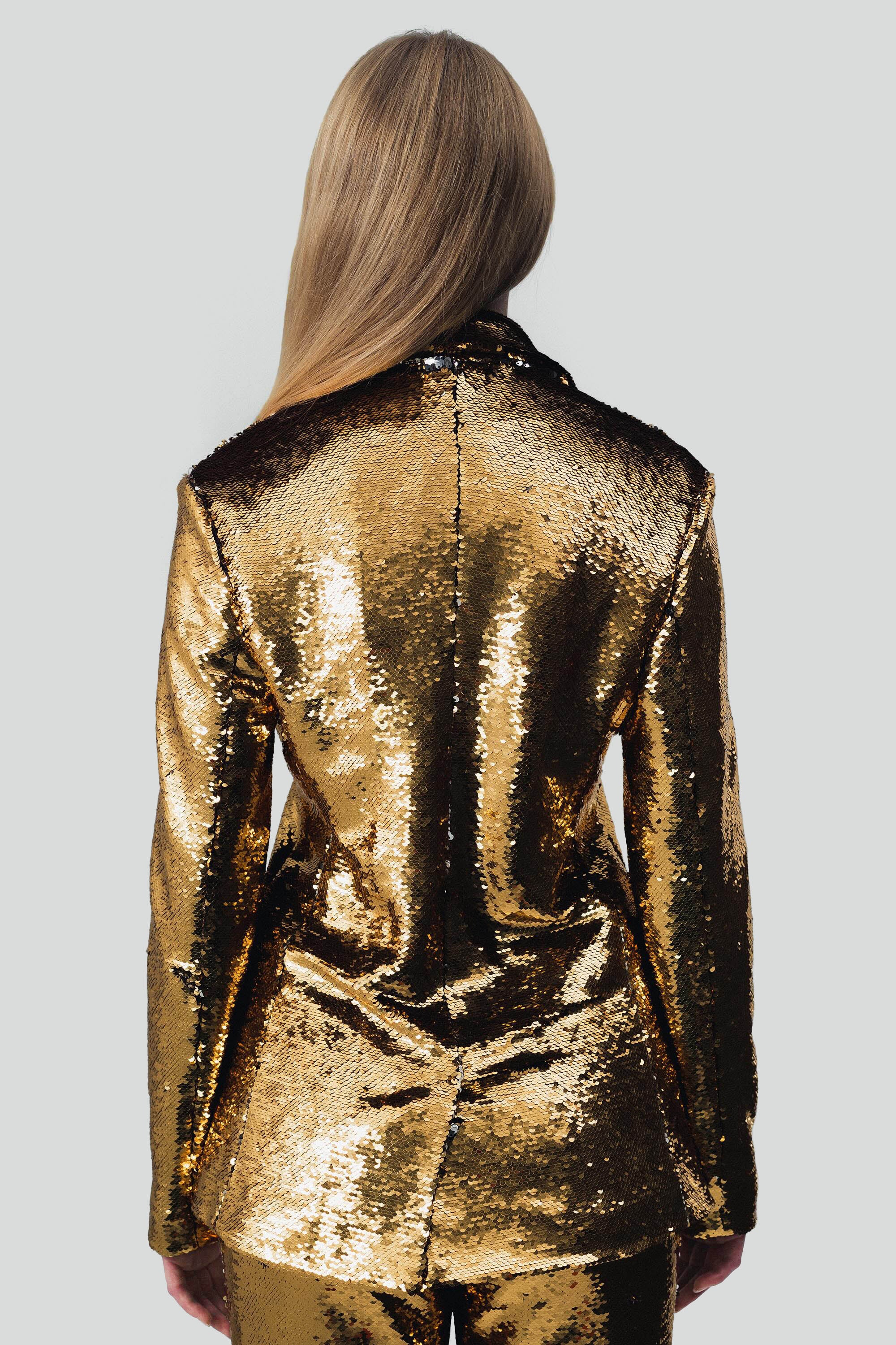 Back view of a woman wearing gold sequin blazer and pants set with a grey background