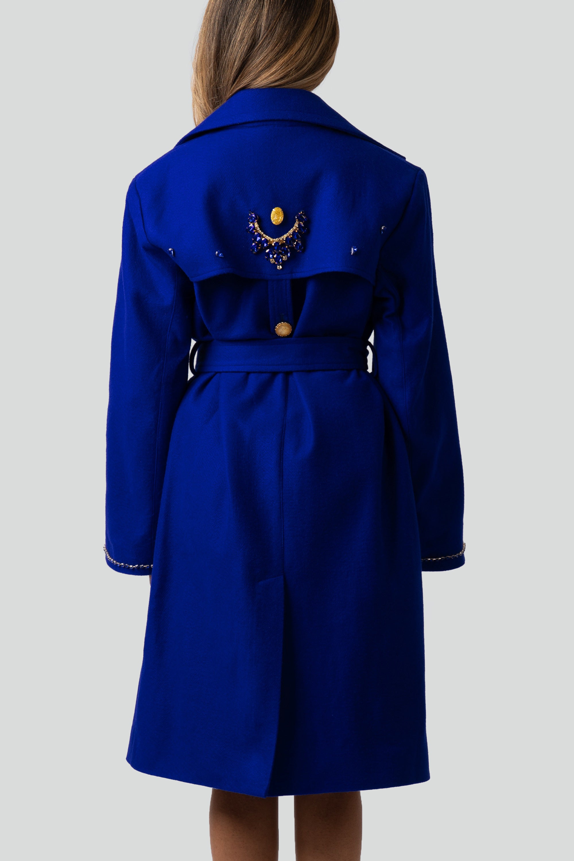 Back of a woman wearing a blue trench coat with gold detailing on a white background