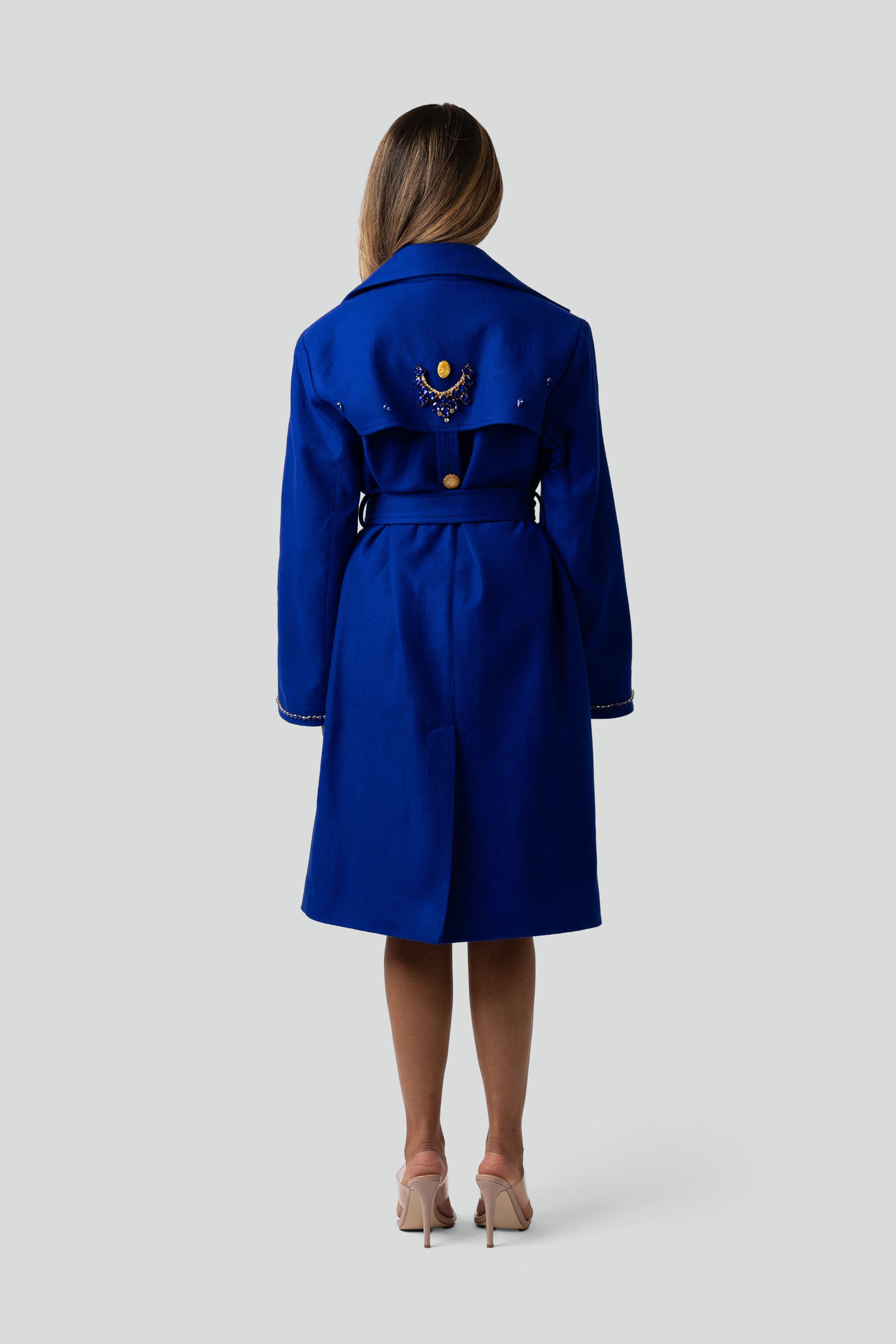 Back of a woman wearing a blue trench coat with gold detailing on a white background