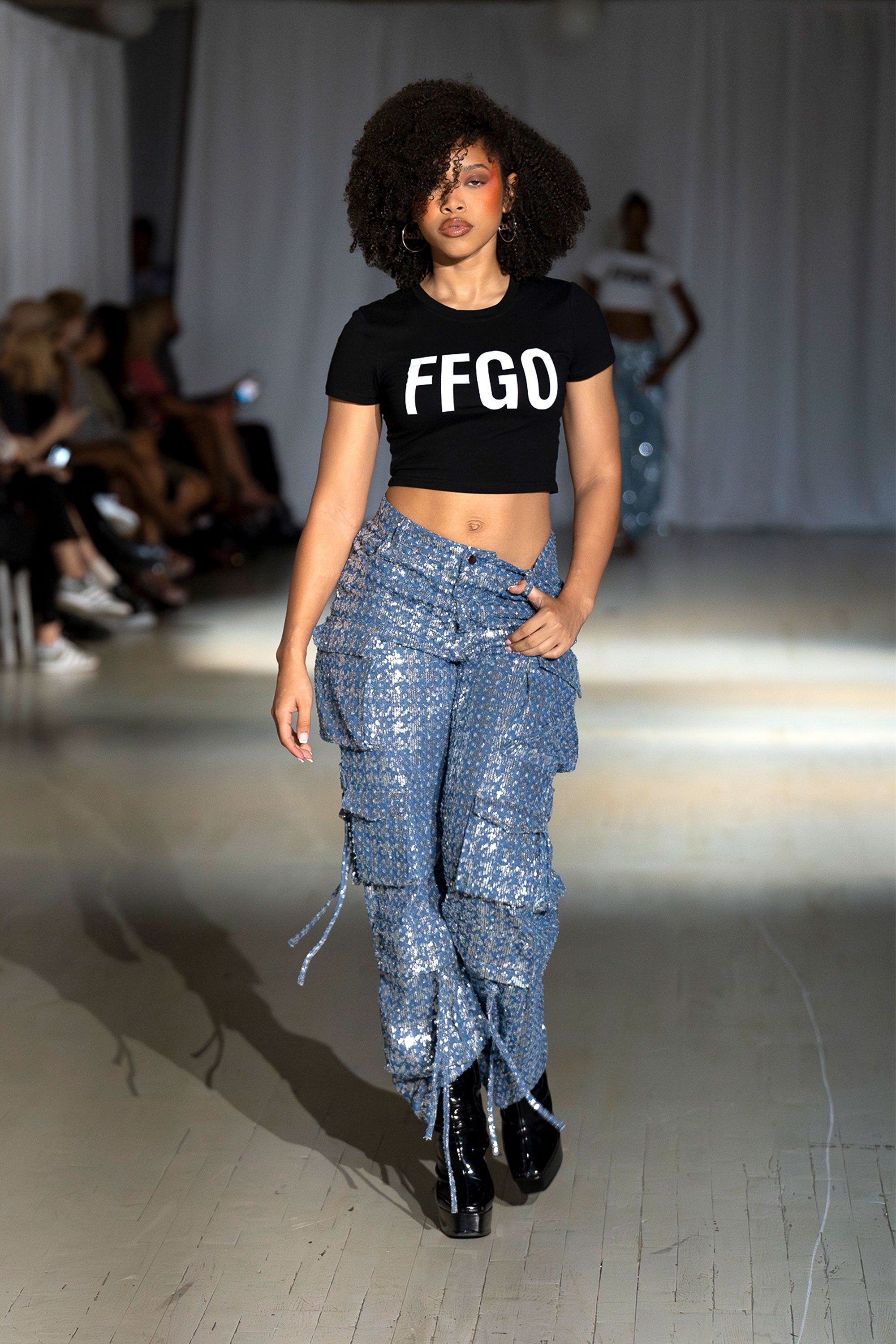 A model walking down a runway wearing blue sequin denim cargo pants with multiple pockets.