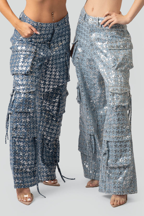 A macro close-up view of premium blue cotton denim fabrication fully embellished with rows of glittering silver sequins, spotlighting a classic zip-fly closure and polished silver button accent