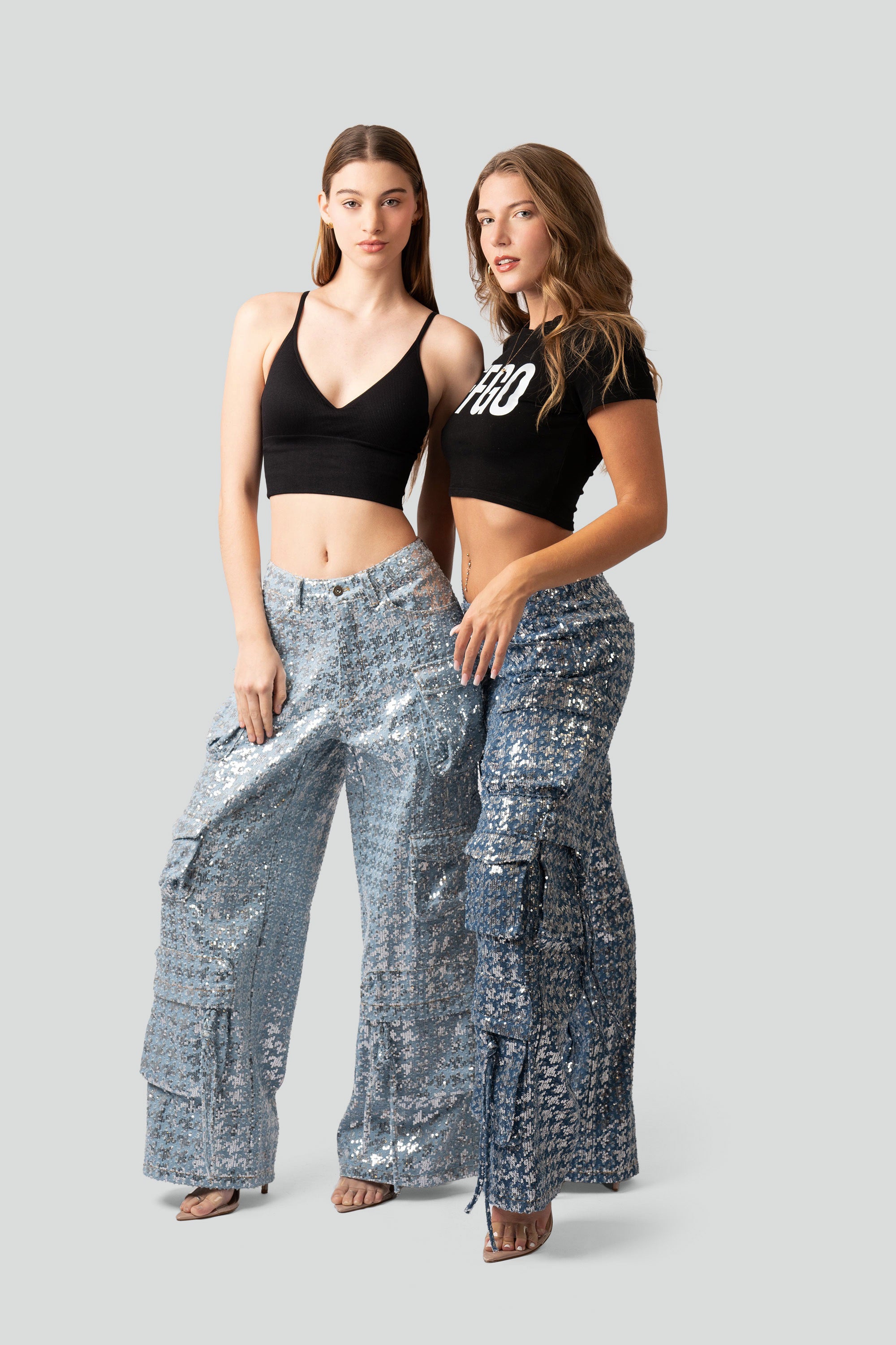 Two women wearing black crop tops and silver sequin pants on a plain background