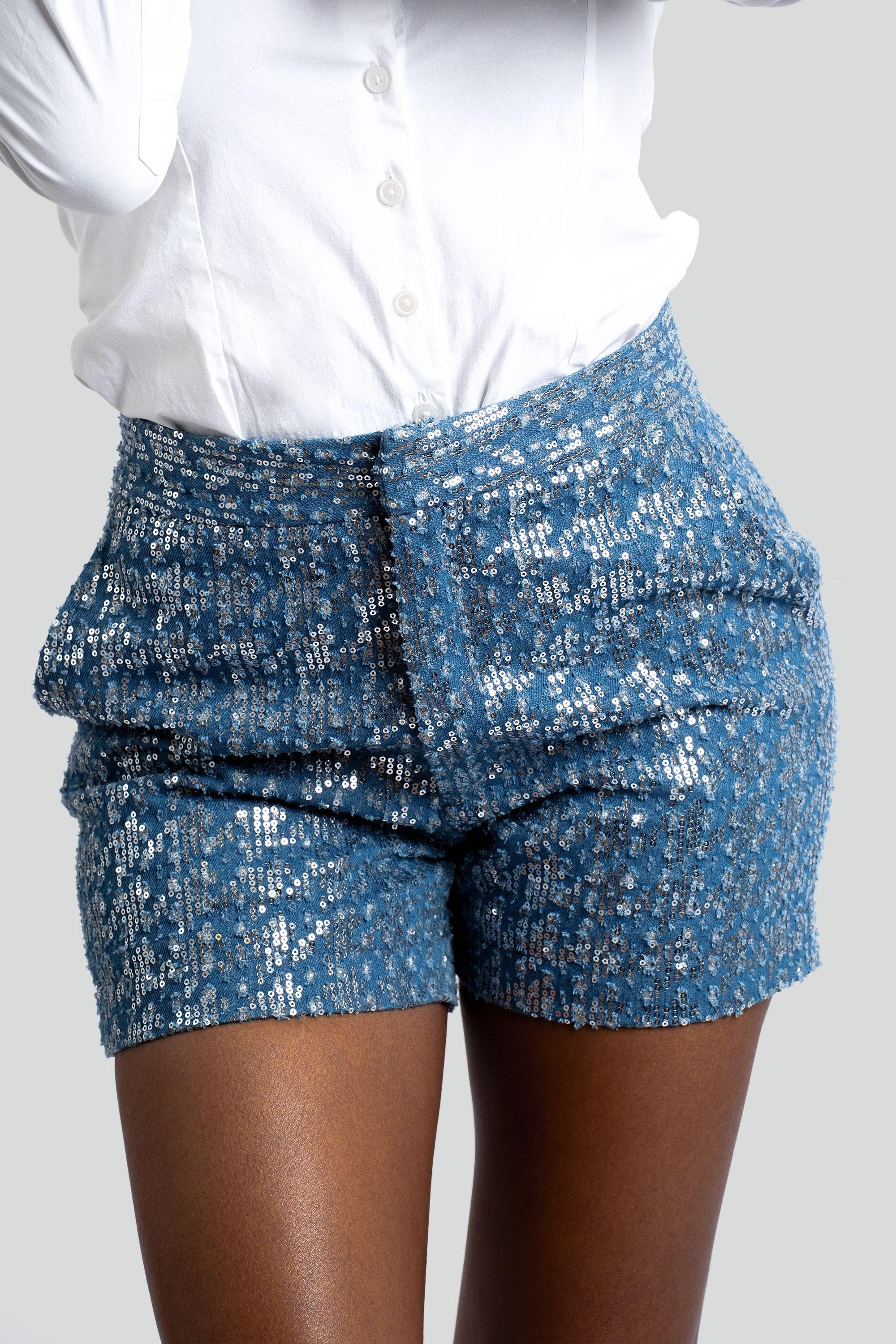 Close up of high-waisted sequined blue denim shorts with a white button top posed against a white background
