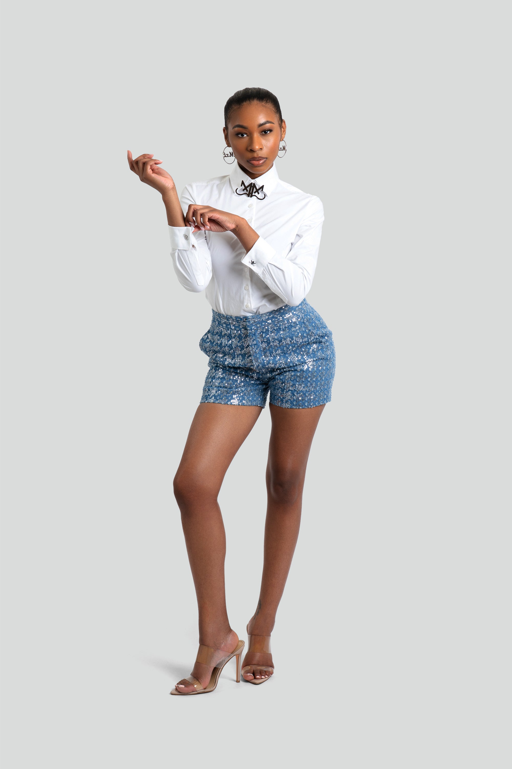 Woman wearing high-waisted blue premium denim shorts covered in micro silver sequins paired with a tailored white button-up shirt by Maui X Lolita