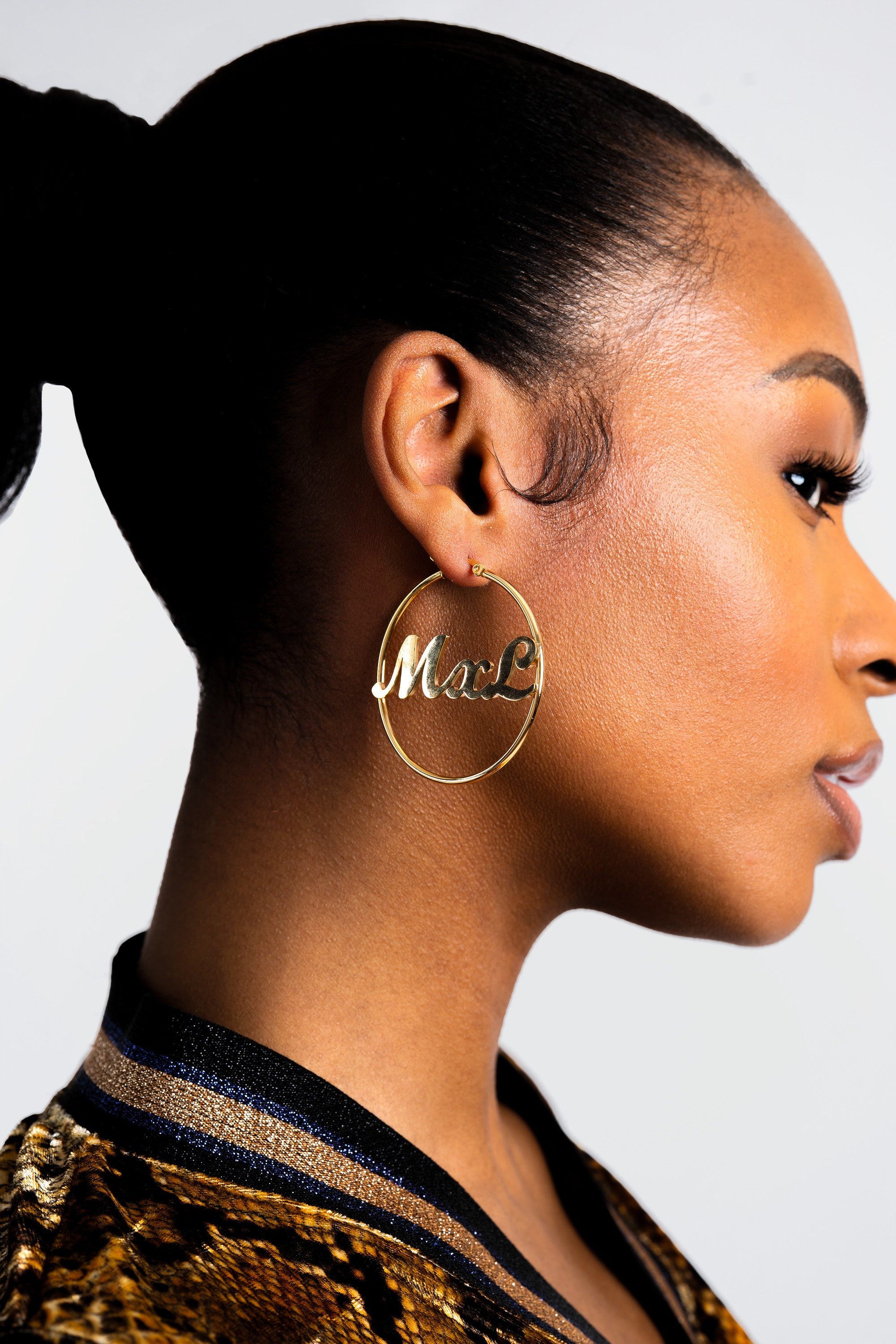 Woman wearing gold hoop earrings with 'MxL' engraving on a plain background