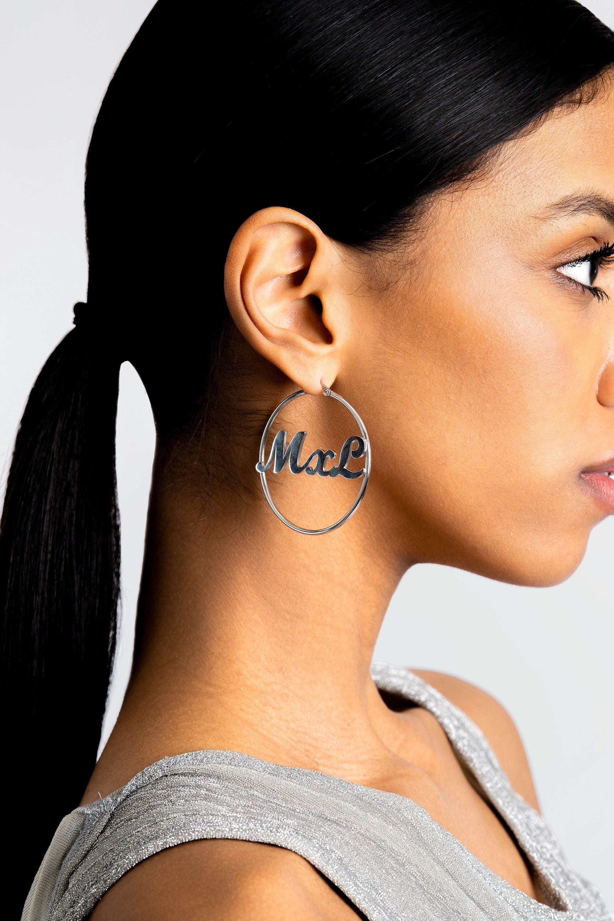 Woman wearing silver hoop earrings with 'MxL' engraving on a plain background