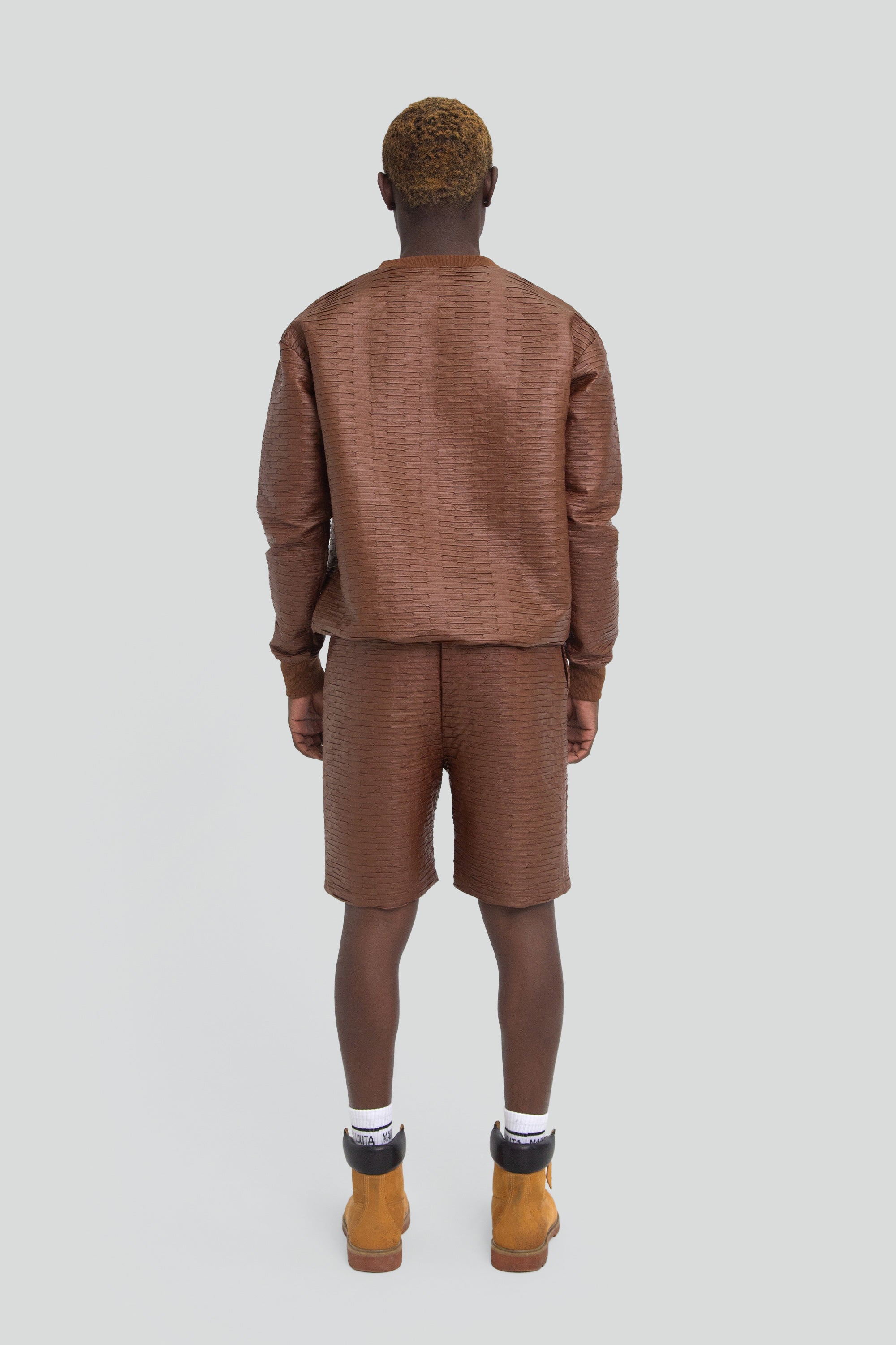 Man wearing a brown textured leather outfit with long sleeves and shorts on a plain background