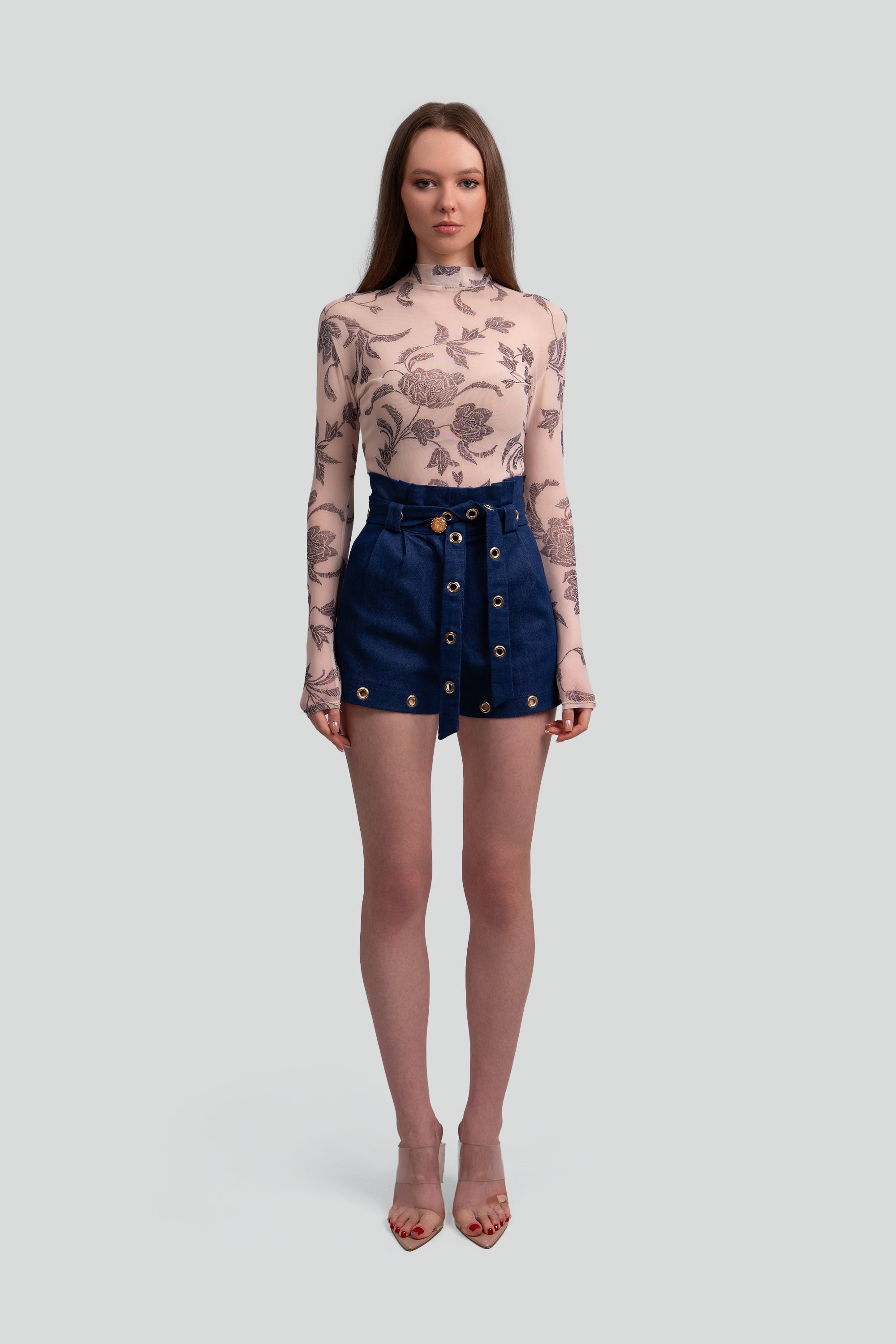 Woman wearing a floral long-sleeve top and navy shorts with gold grommets on a white background