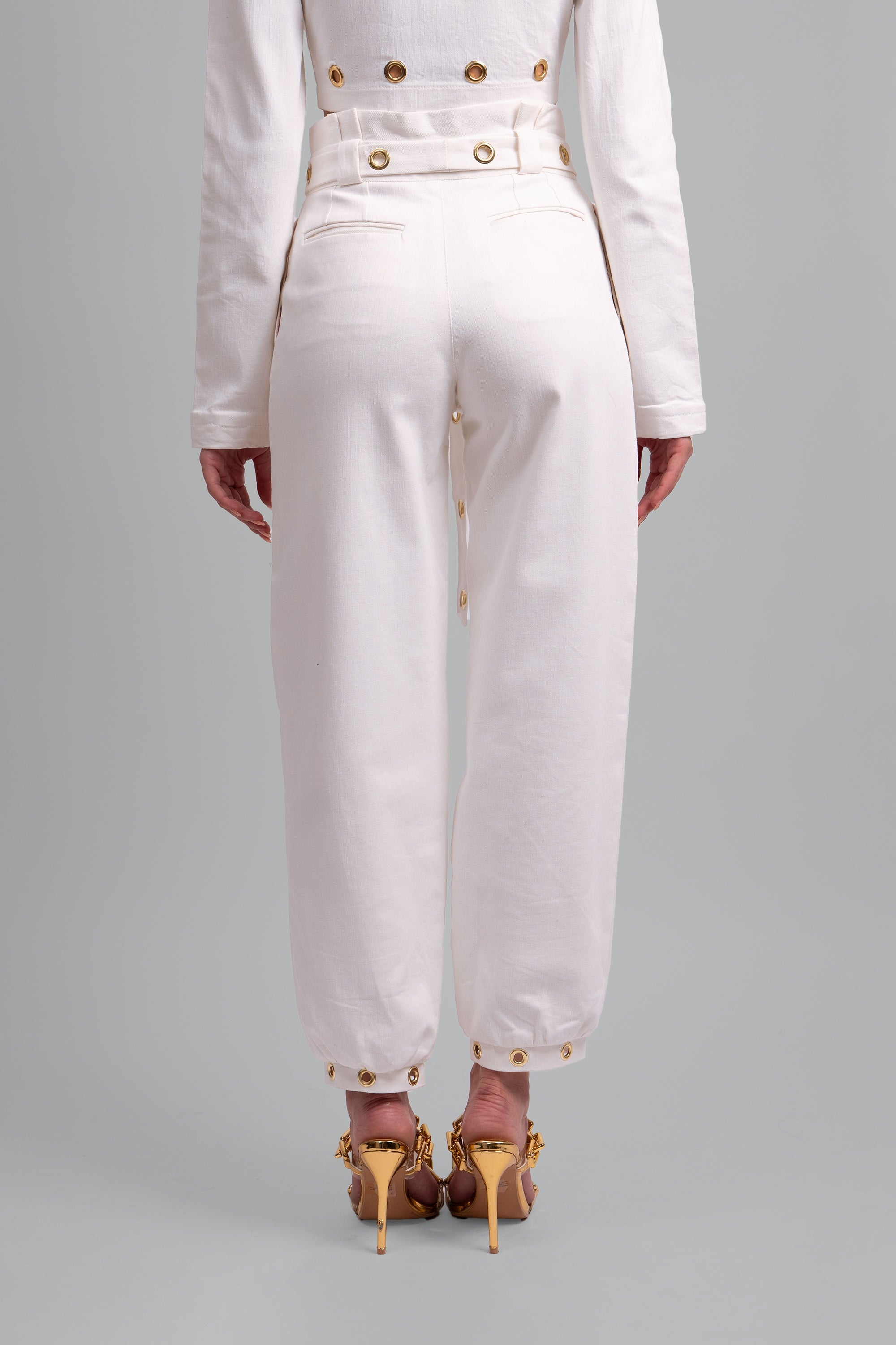 Back view of a woman wearing a white pant with gold grommet details on a gray background