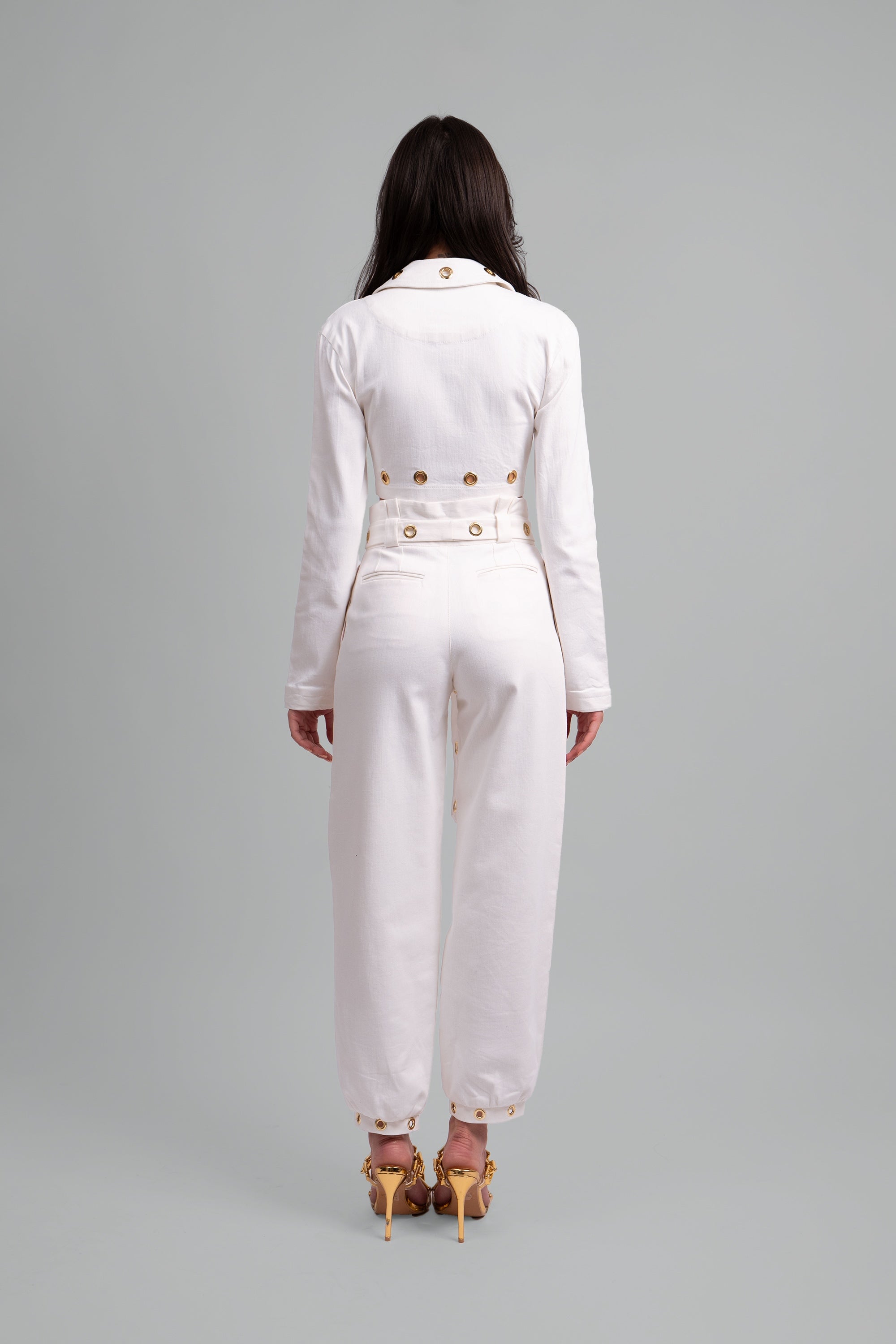 Back view of a woman wearing a white two piece cropped set with gold grommet details on a gray background