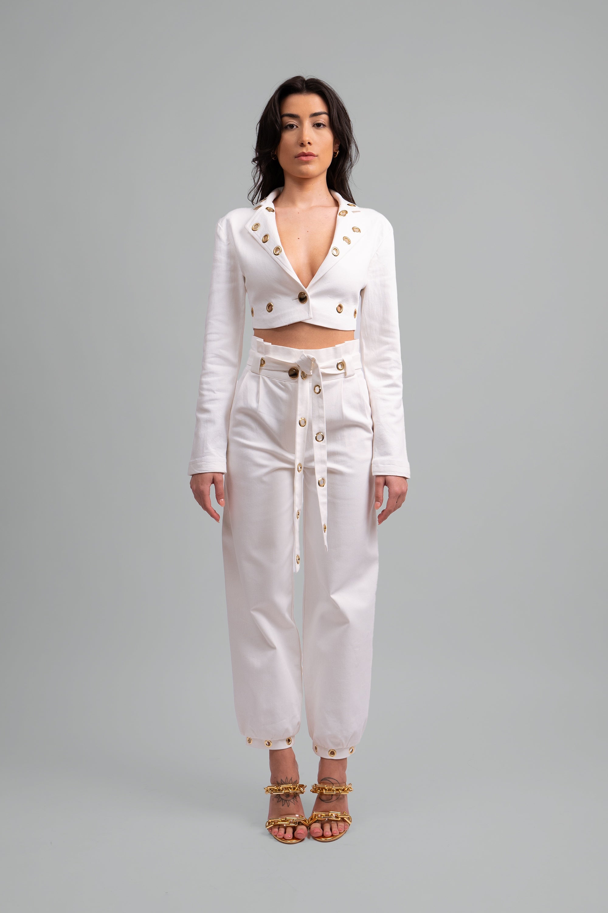 Woman wearing a white two piece cropped set with gold grommet details on a gray background