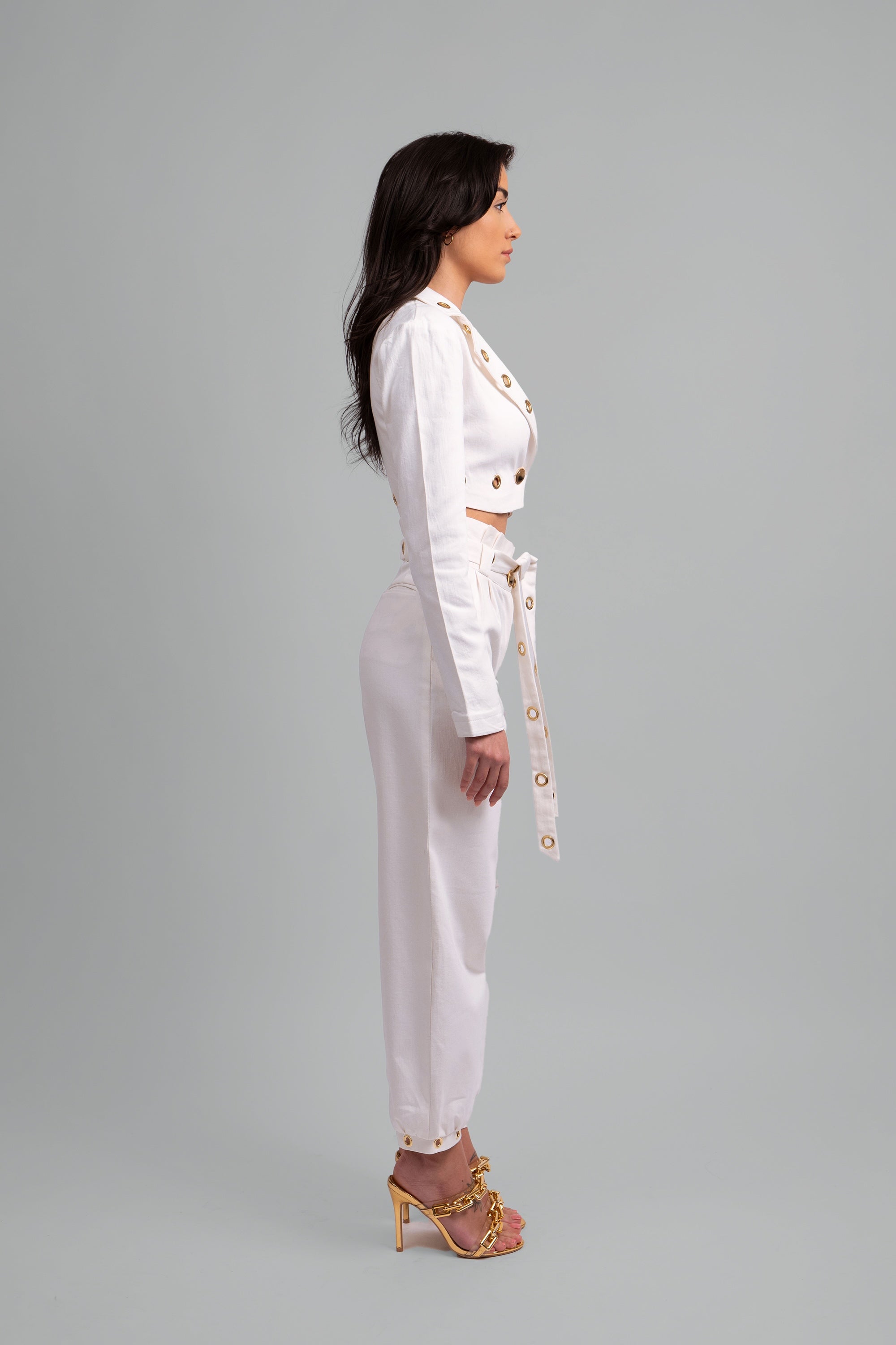 Side view of a woman wearing a white two piece cropped set with gold grommet details on a gray background