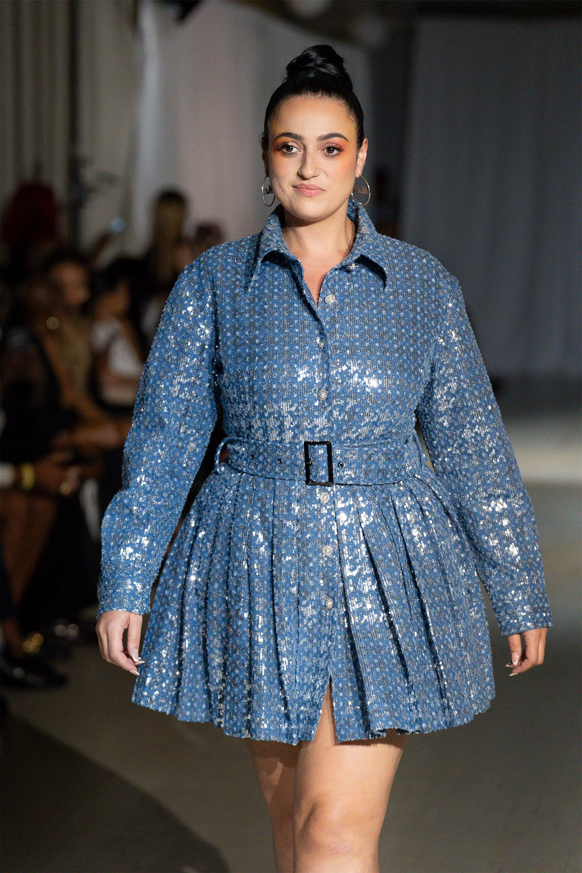 A woman modeling a blue denim mini dress with silver sequins and pearl crystal buttons, featuring a tailored button-up bodice, a sharp collar, a voluminous pleated skirt, and a waist cinch belt.
