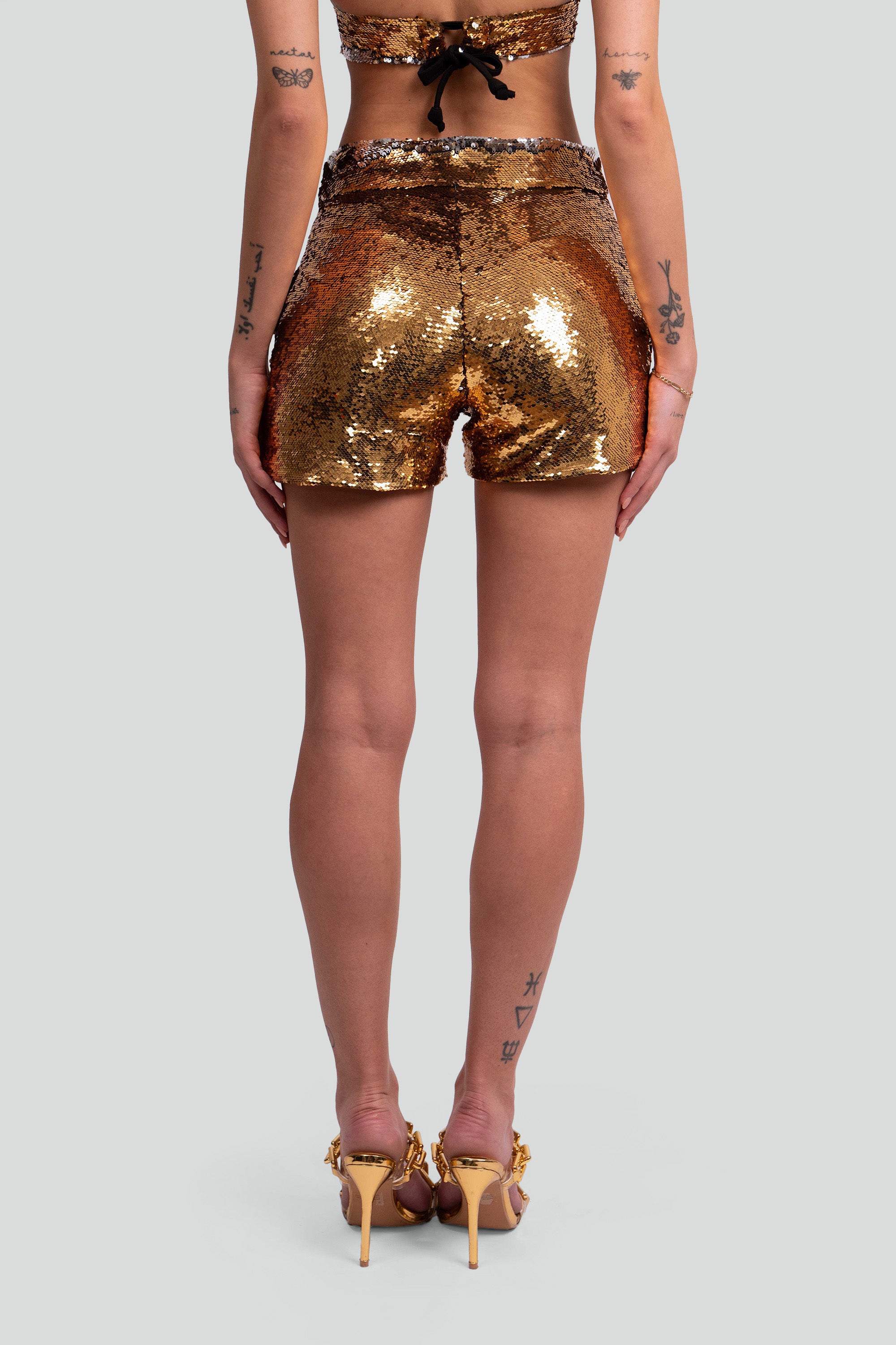 Back view of a woman wearing gold sequin shorts on a white background