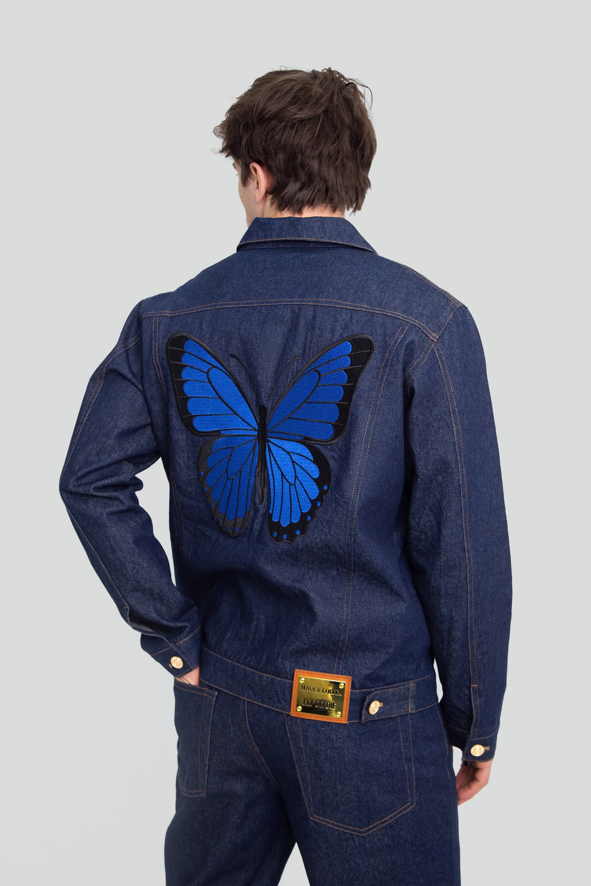 Person wearing a blue denim jacket with a large blue butterfly design on the back against a plain background