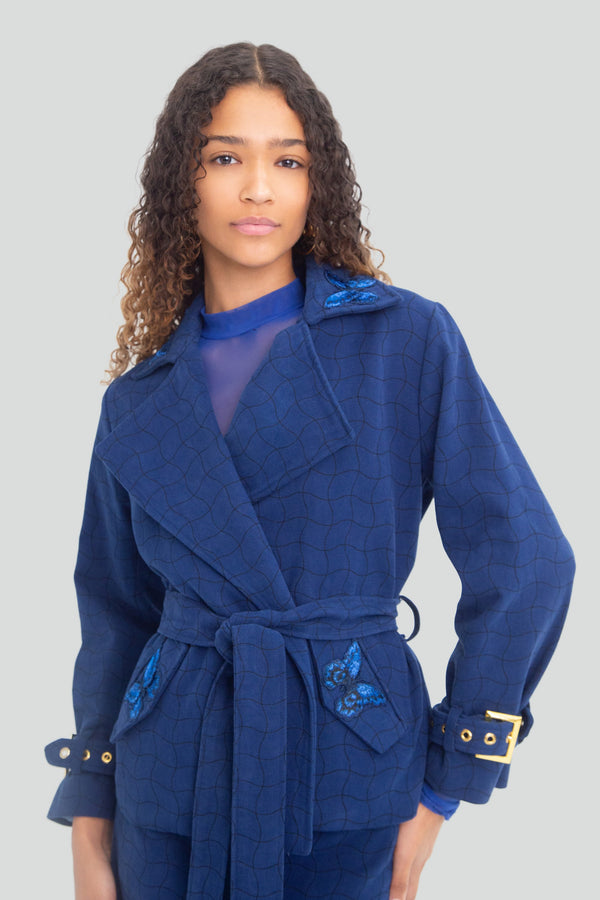 Woman wearing a light blue suede like denim cropped trench jacket featuring an adjustable waist belt and butterfly pocket details