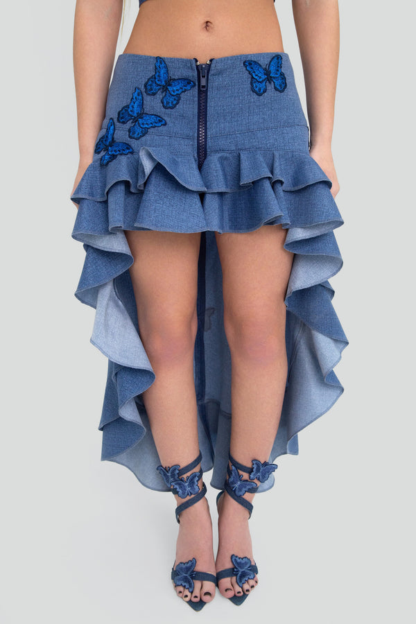 Woman wearing a blue medium wash denim high low skirt featuring a low waisted fit and dramatic ruffled silhouette
