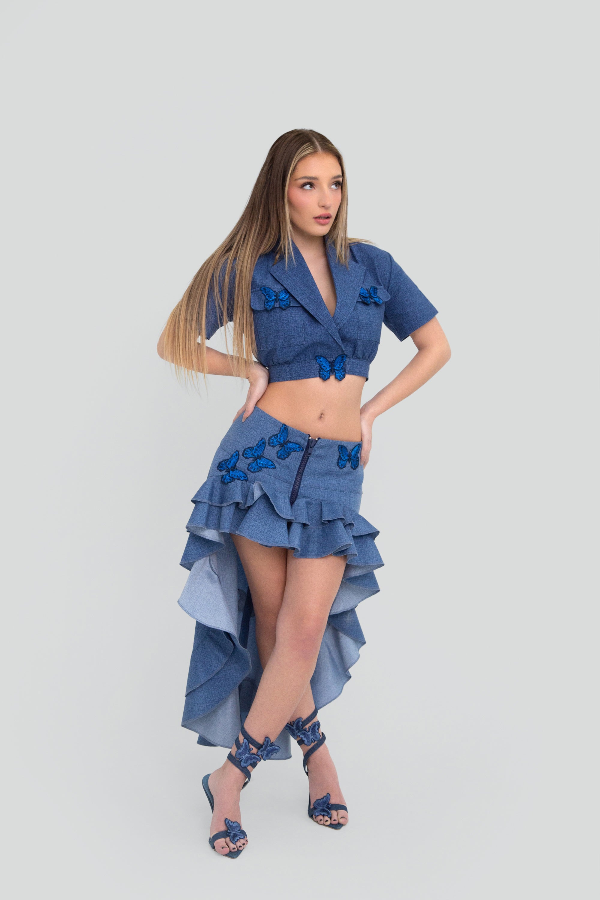 Woman wearing a blue outfit with a high-low ruffled skirt and butterfly details on a plain background