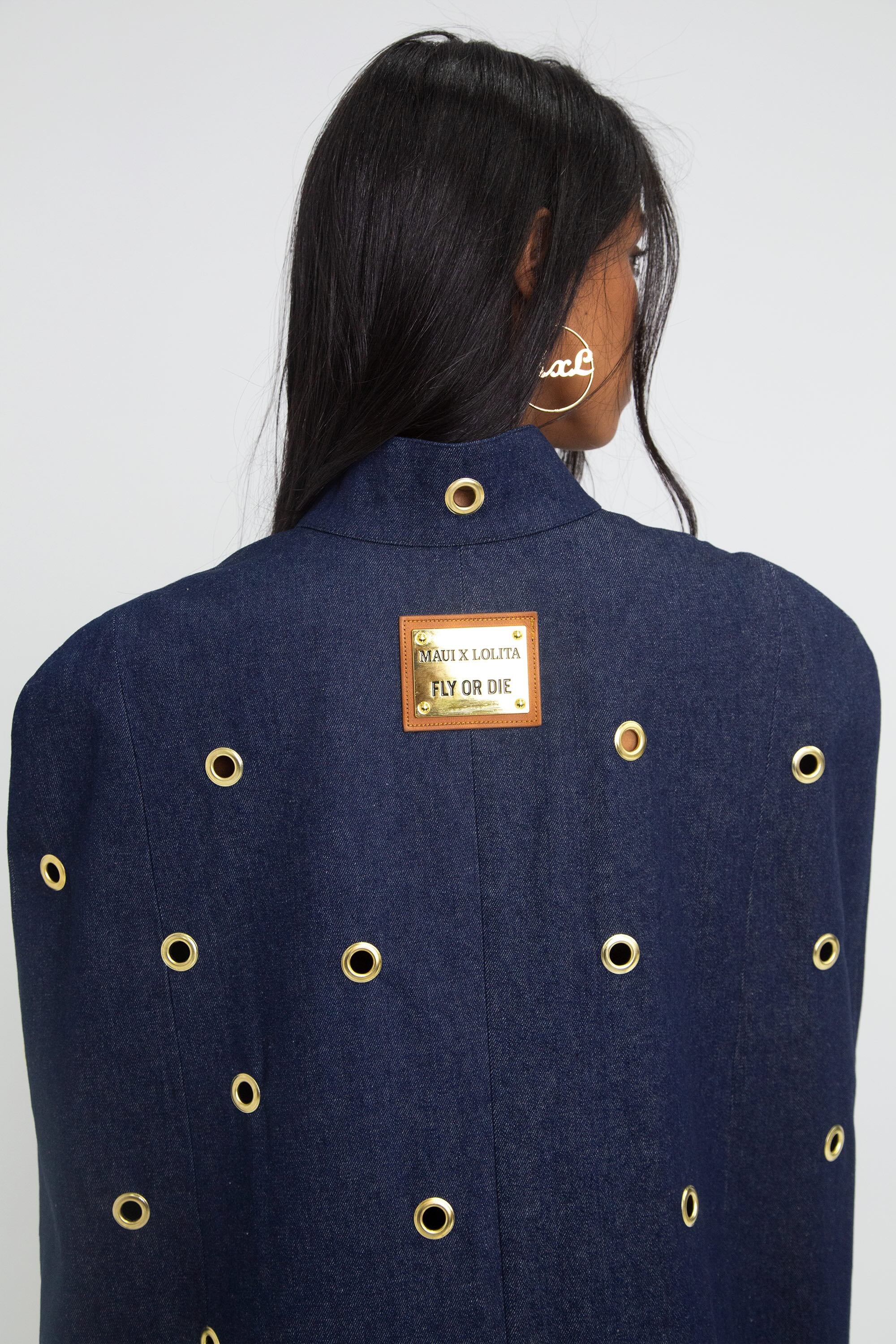 Close up of a woman wearing a dark blue denim cape with gold grommet embellishments on a plain background