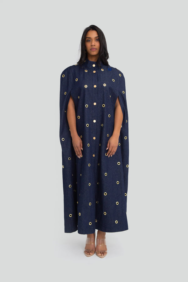 Woman wearing a full length deep indigo denim cape featuring a structured high collar, gold button details, and a bold front leg slit