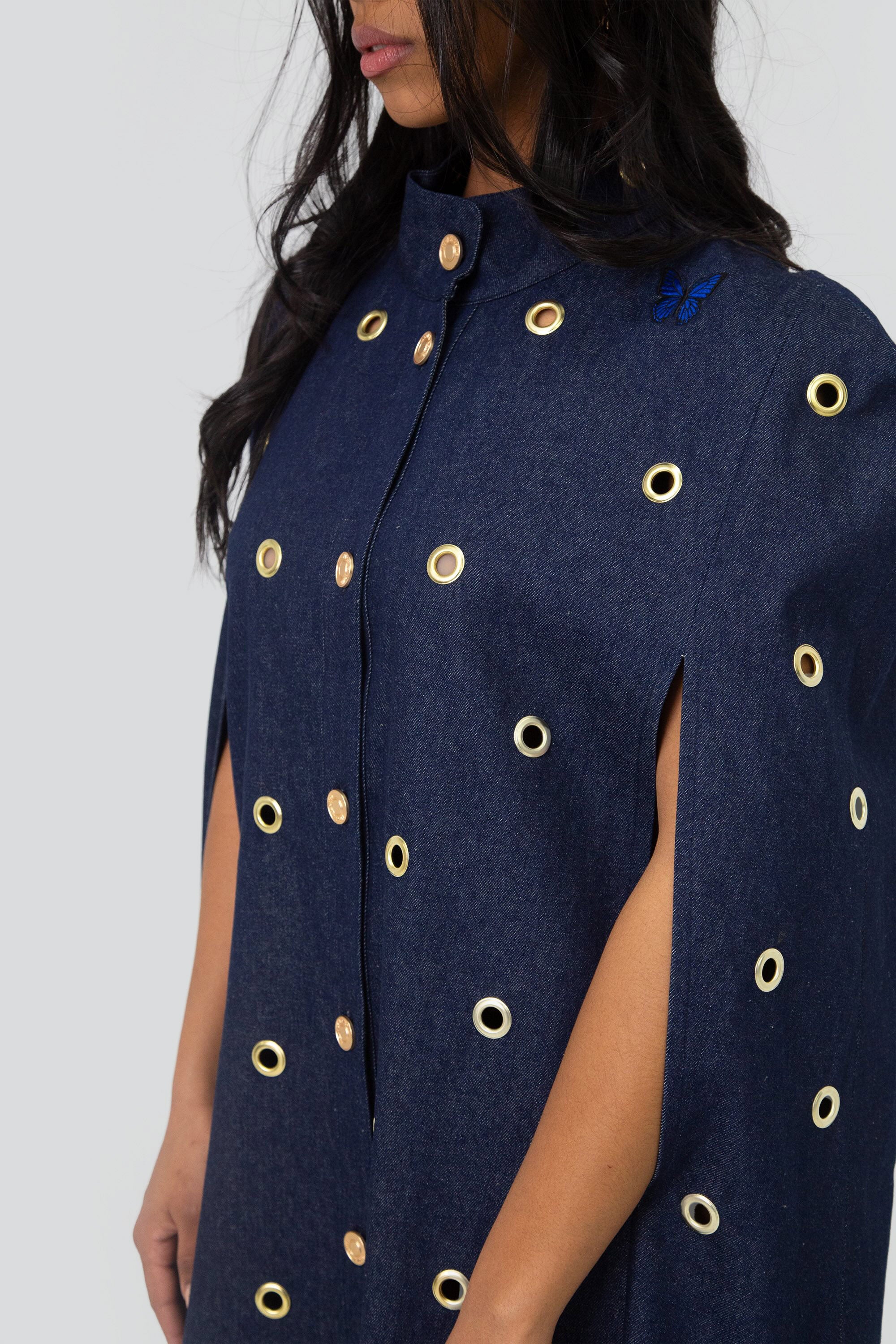 Close up of a woman wearing a dark blue denim cape with gold grommet embellishments on a plain background
