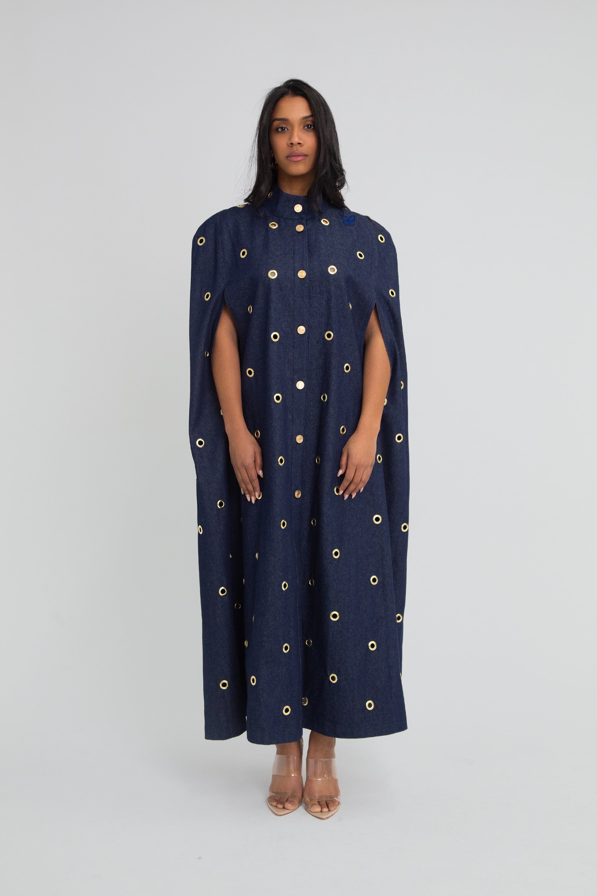 Woman wearing a dark blue denim cape with gold grommet embellishments on a plain background