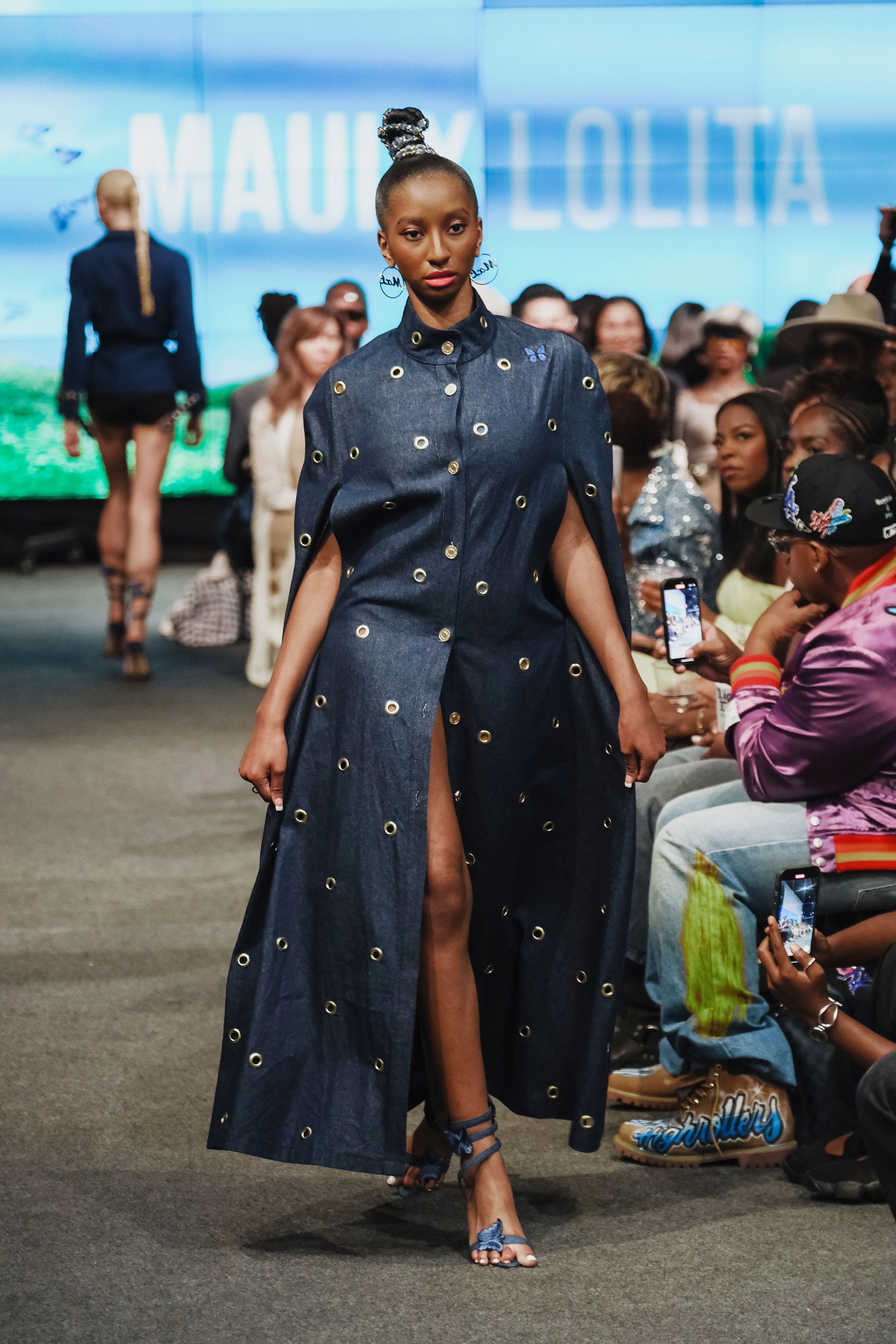 Model walking on a runway wearing a dark blue dress with gold embellishments, with 'MAUI X LOLITA' branding in the background.