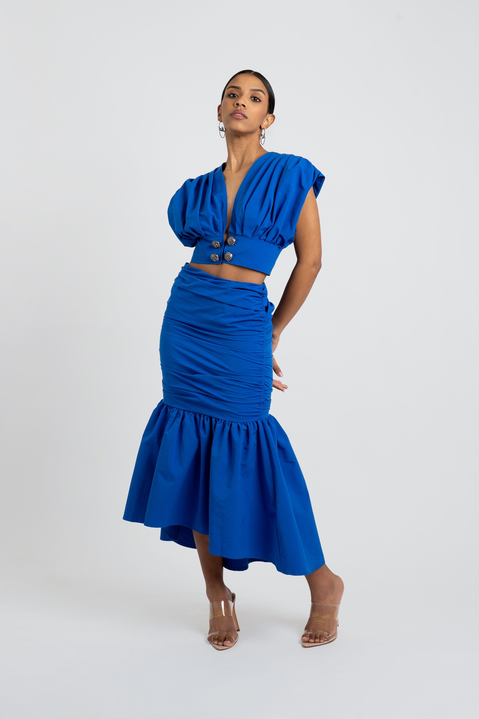 Woman wearing a blue ruched two piece skirt set with silver buttons on a white background