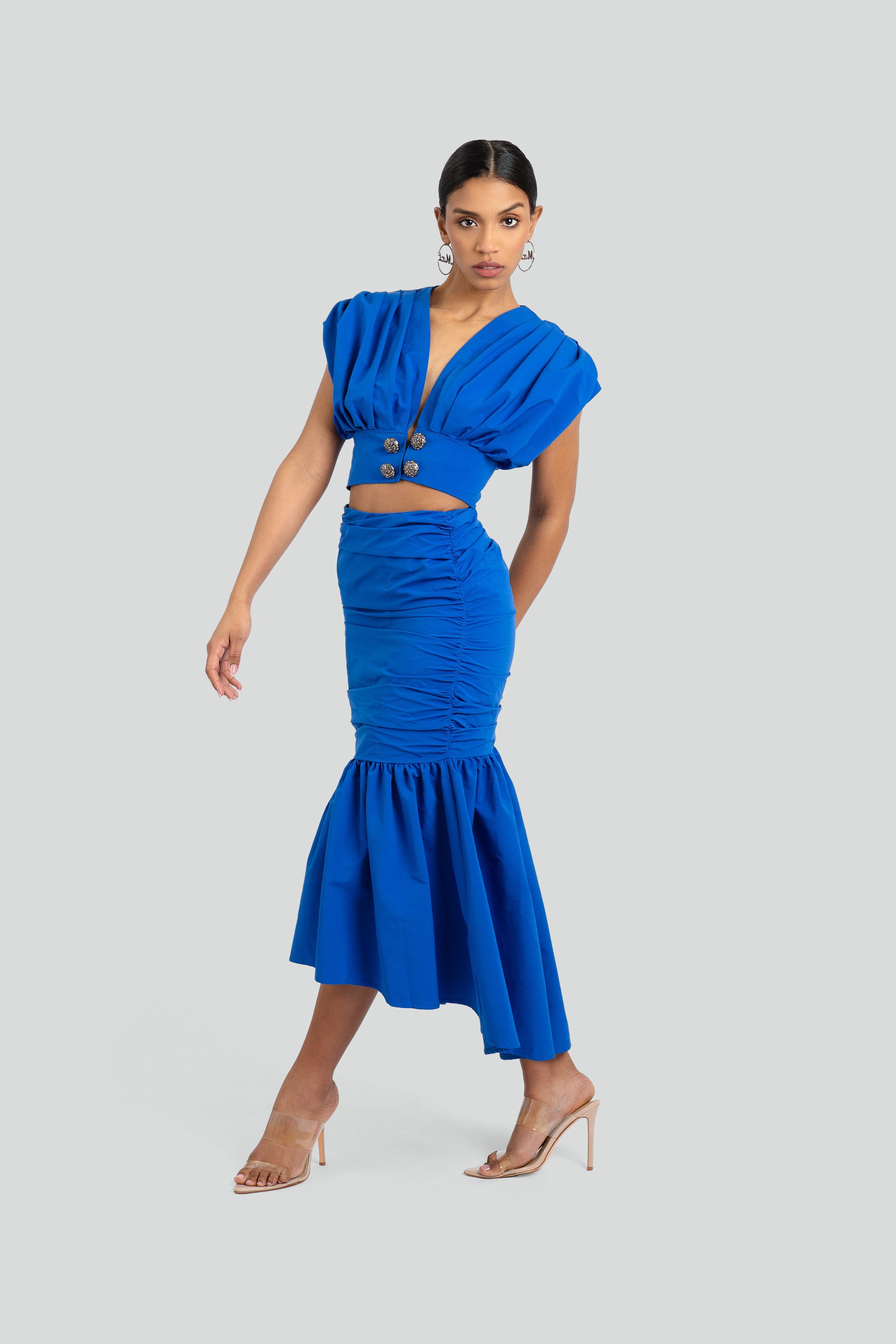 Woman wearing a blue ruched two piece skirt set with silver buttons on a white background