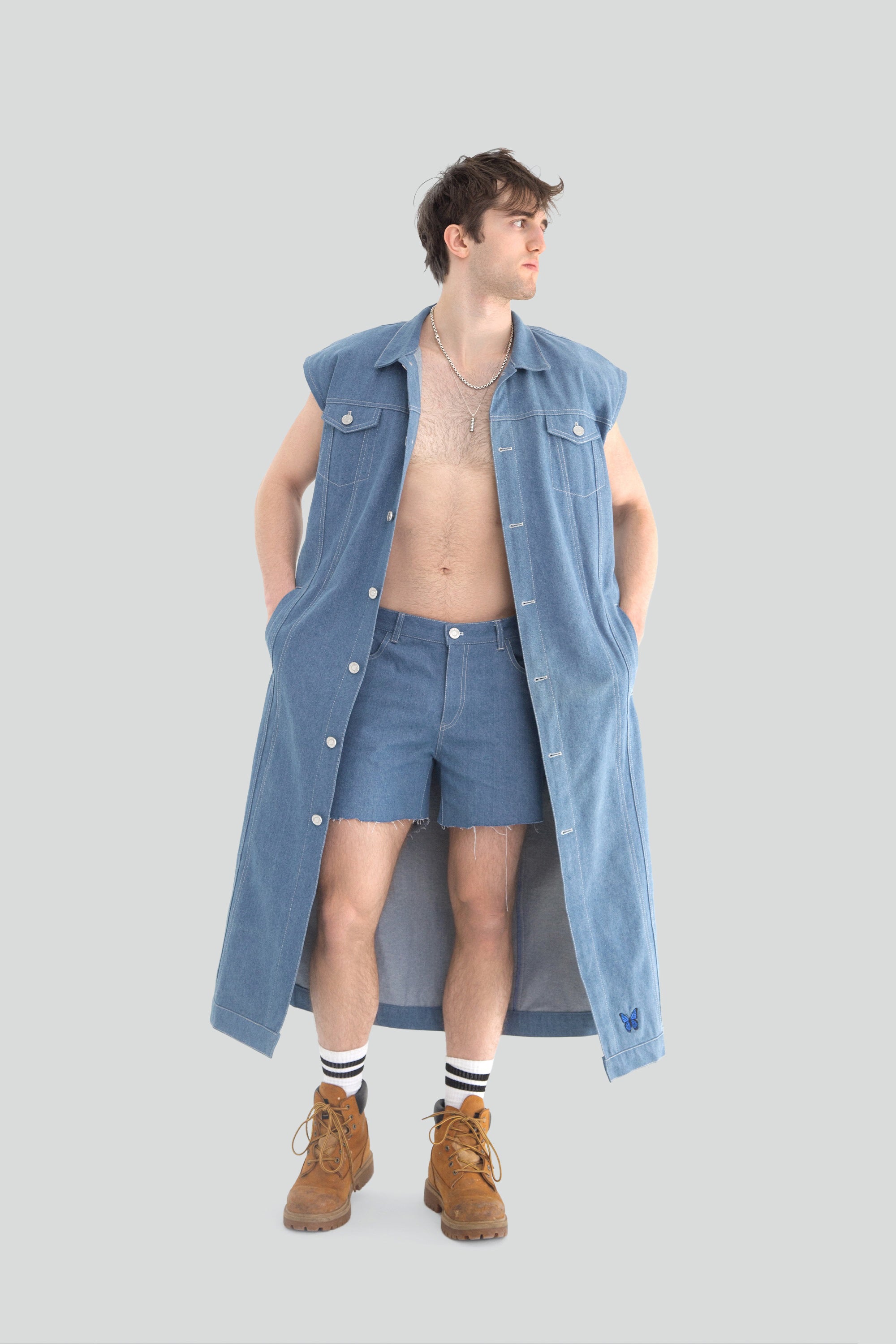 Person wearing a long blue denim vest over shorts on a plain background