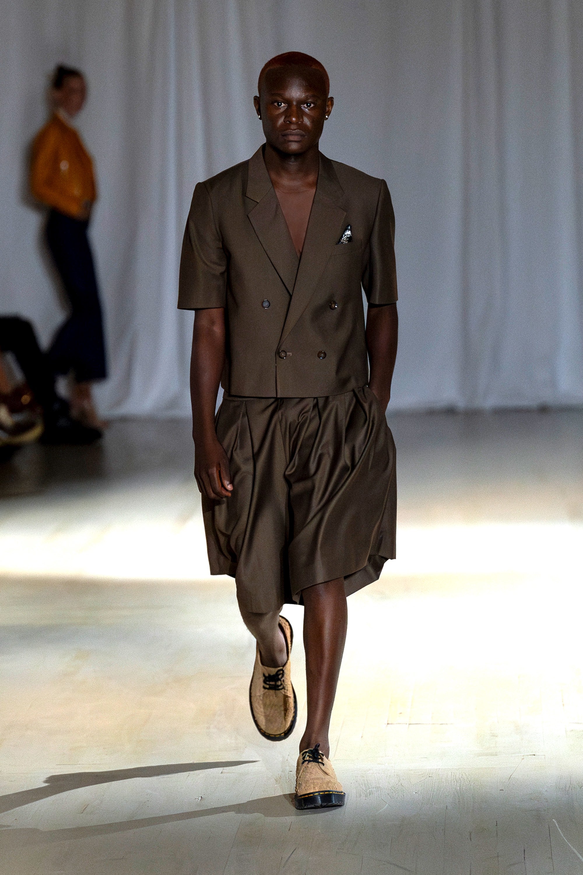 Model wearing a cropped brown blazer and short set walking down the runway.