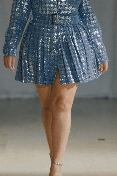 Model wearing a blue sequined dress with a sharp collar, belt, and pleated skirt walking down the runway.