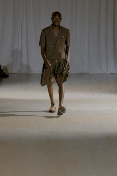 Model wearing a cropped brown blazer and short set walking down the runway.