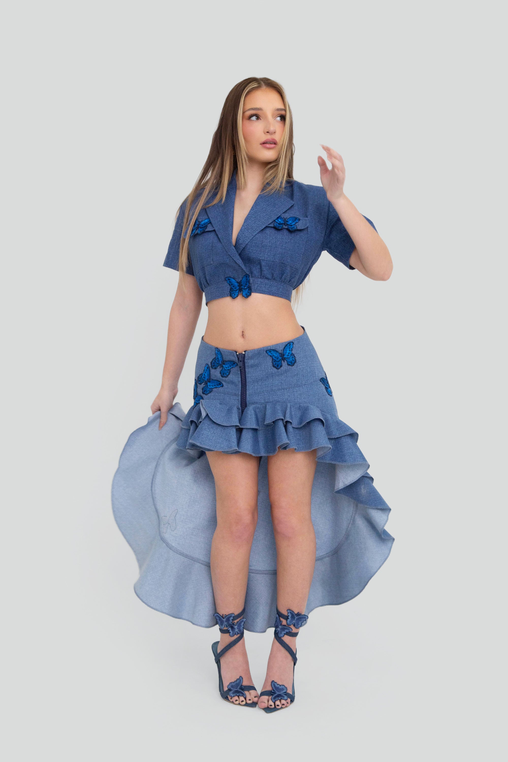 Woman wearing a blue outfit with a high-low ruffled skirt and butterfly details on a plain background