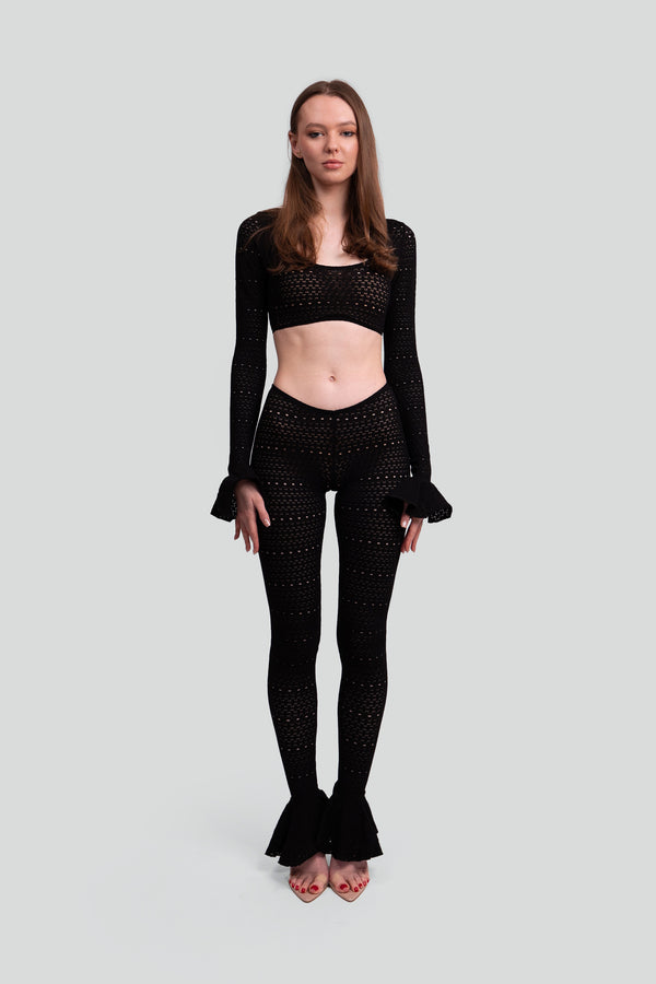 Woman wearing a black lace knit matching set featuring a ruffled long sleeve crop top and low rise sheer pants